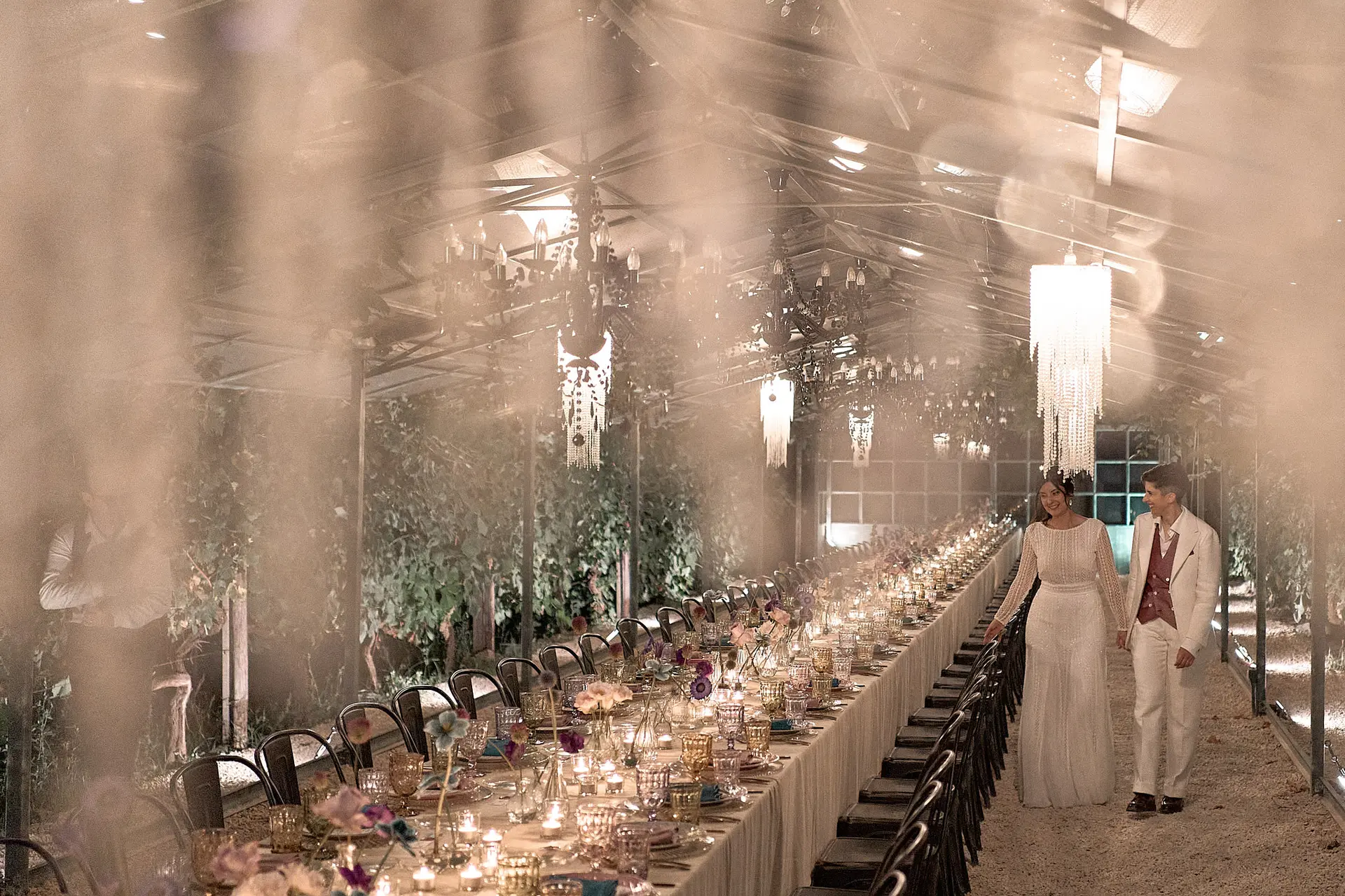 Two brides walk a long table, greenhouse setting. A stylish, atmospheric vegan wedding reception.
