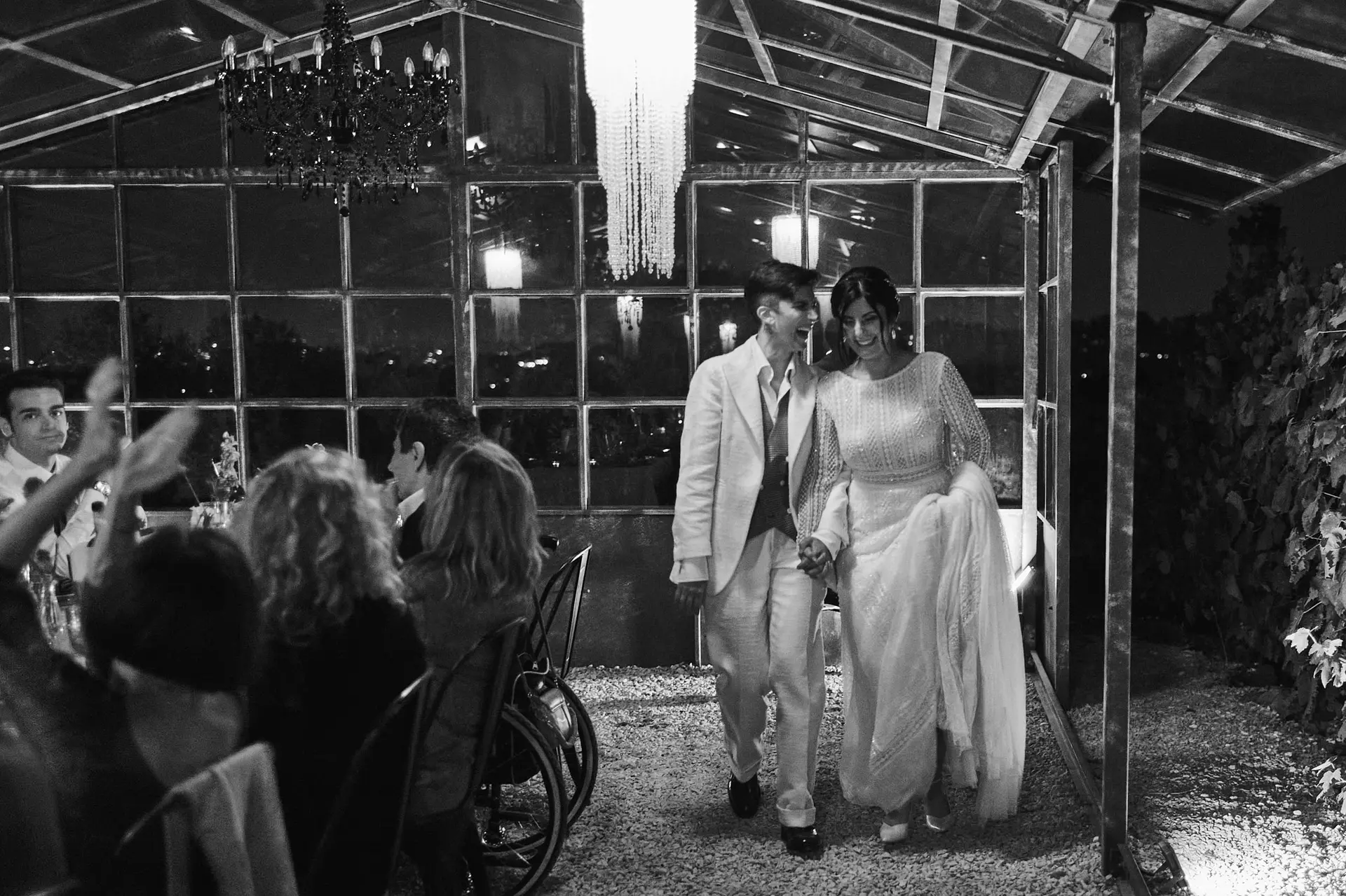 Monochrome photo, two brides walk hand-in-hand entering between people is like a runway.