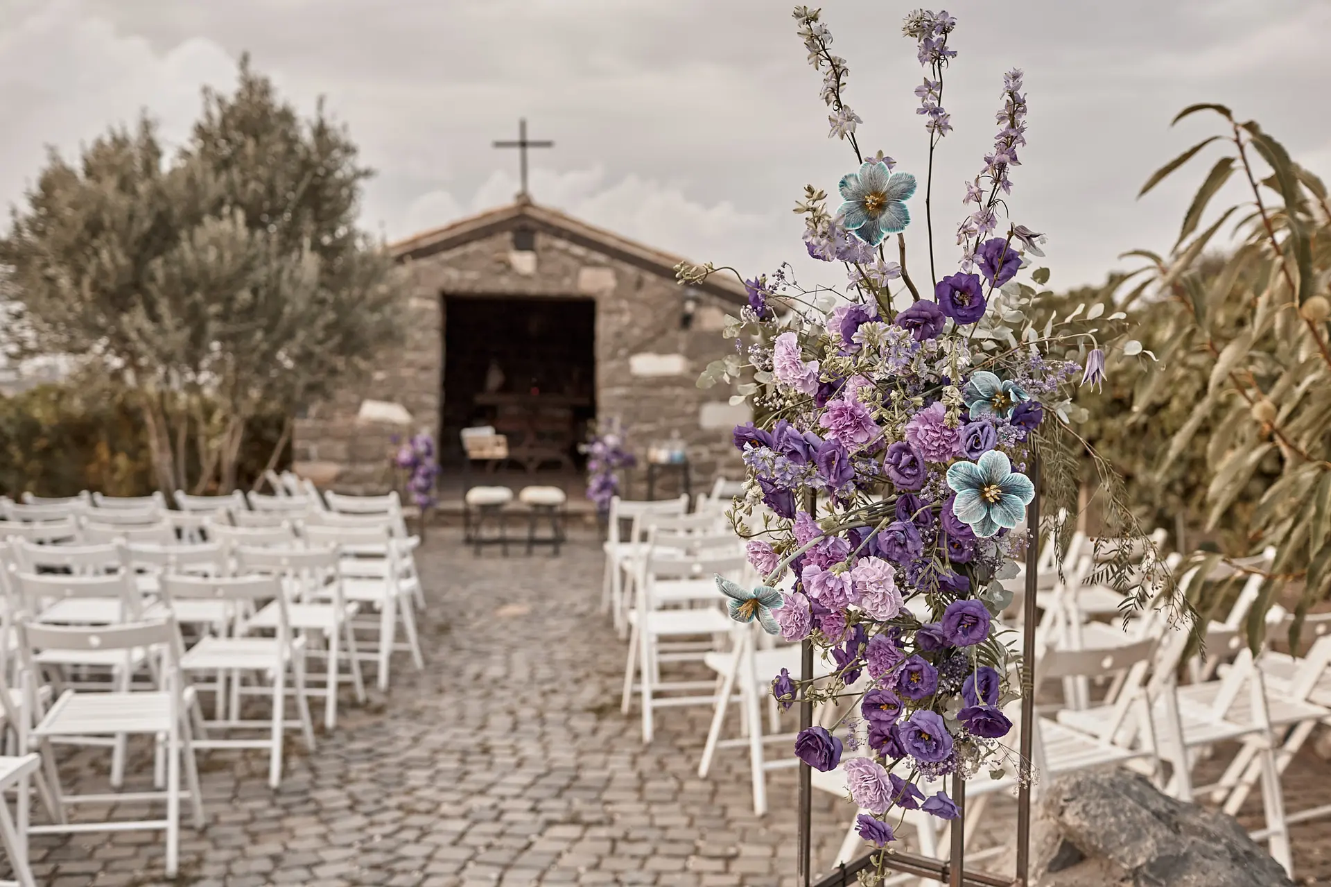 Outdoor wedding rehearsal aisle with purple flower arrangement leading towards stone chapel under cloudy sky.