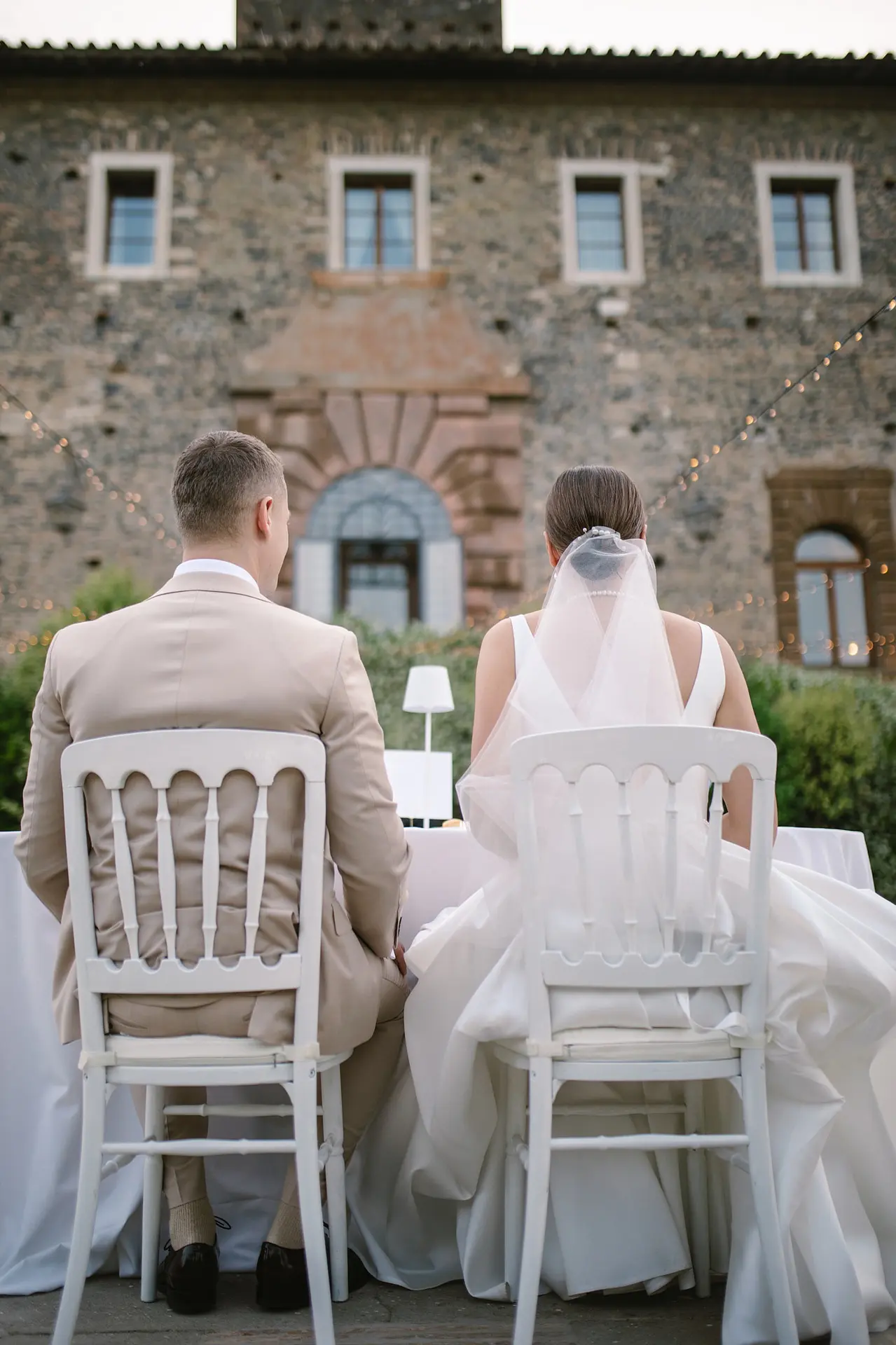 Wedding rehearsal dinner setup in historic location, bride and groom sitting at table facing building.