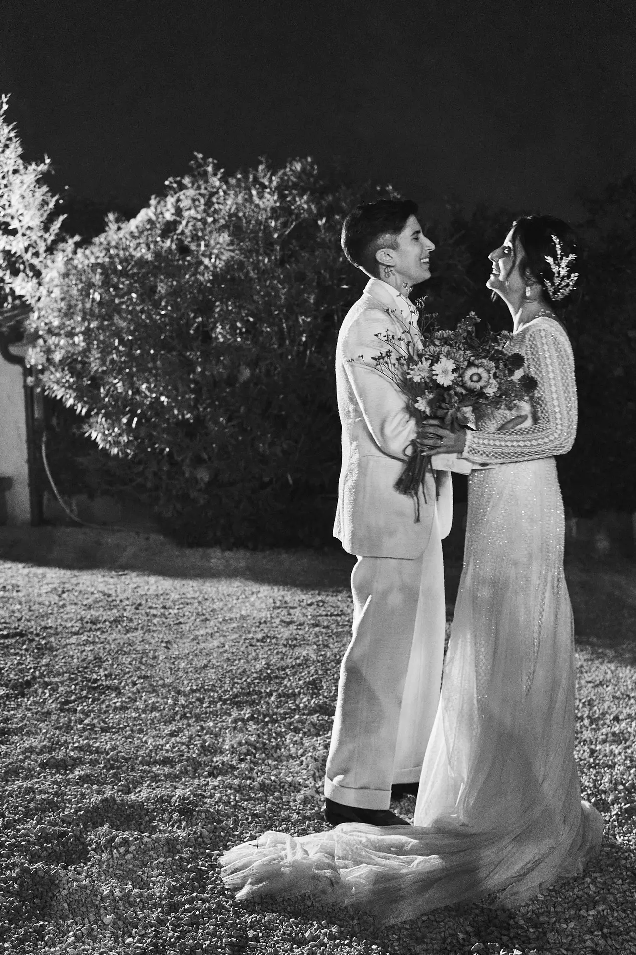 Two brides lovingly embrace at their outdoor nighttime wedding rehearsal in garden setting.