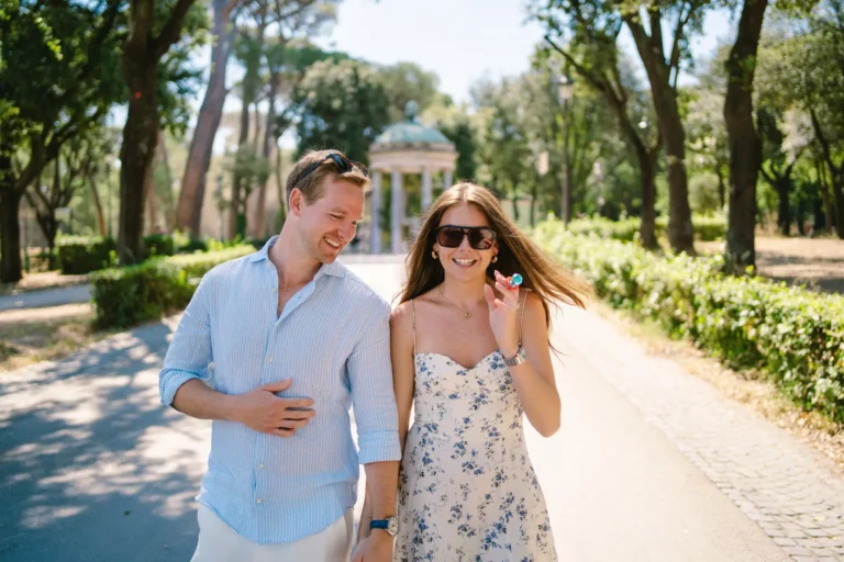 Happy couple walks hand-in-hand in a park, woman shows ring box.