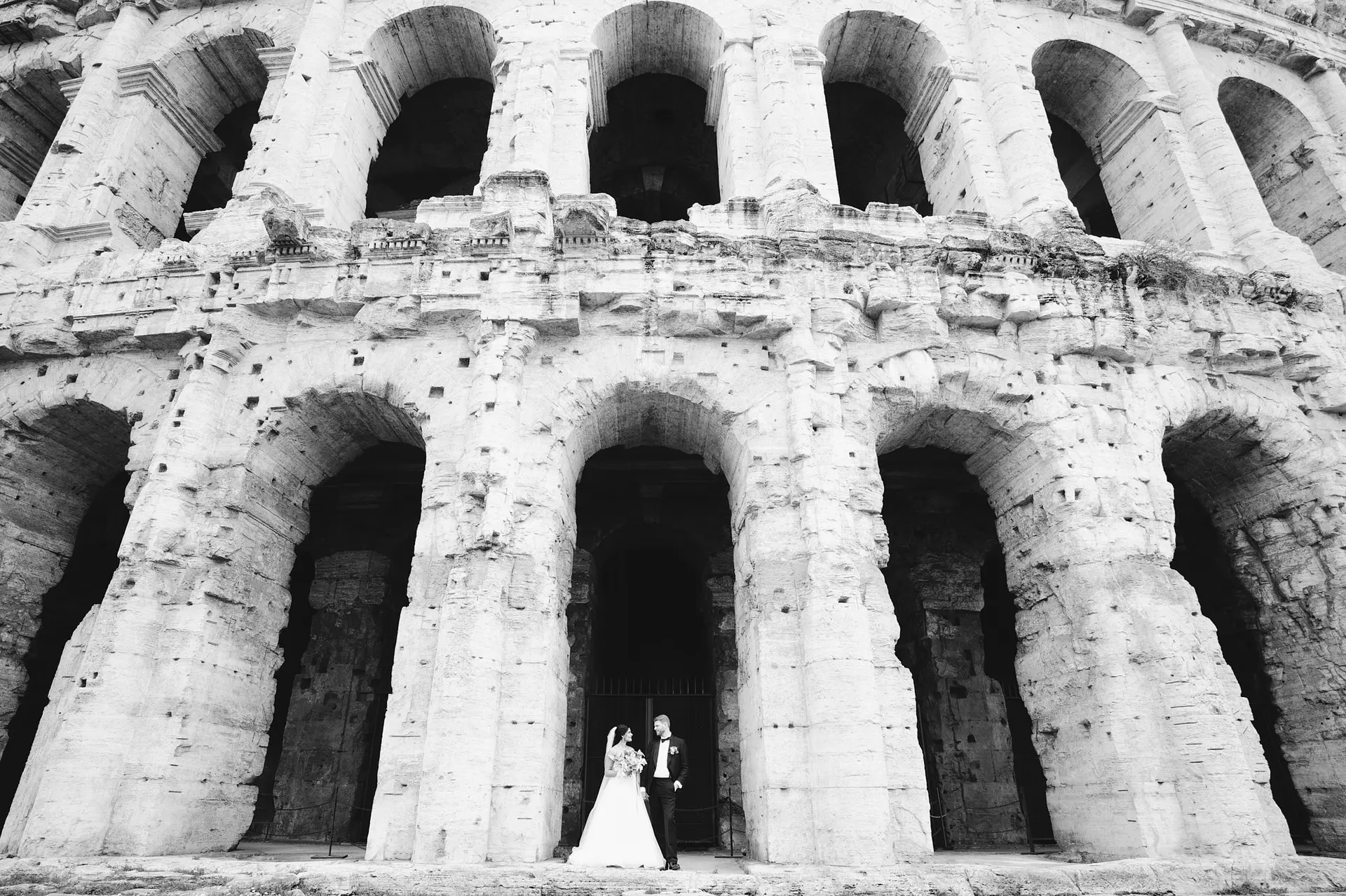 B&W: Tiny wedding couple stands in front of the massive arches of Teatro Marcello.