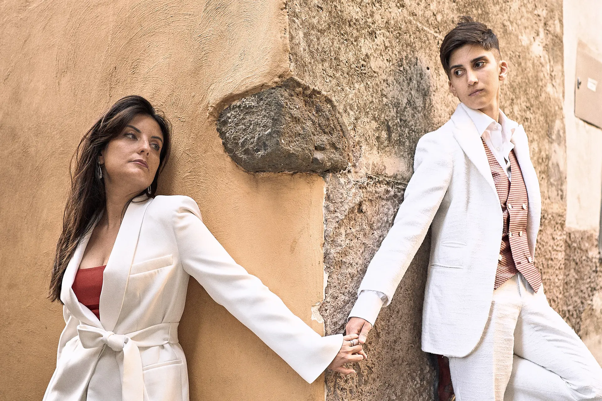 Two women in white suits hold hands around the corner of a tan wall.