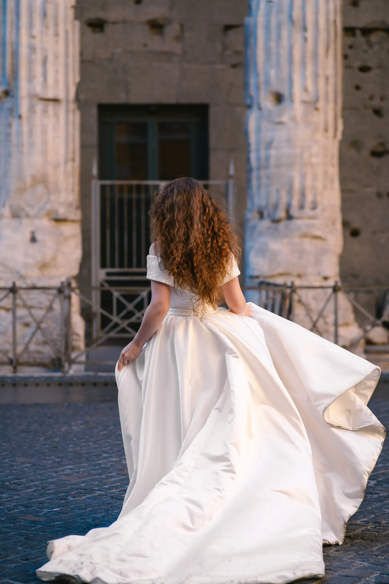 Bride with long curly hair walks away in a flowing white wedding dress.