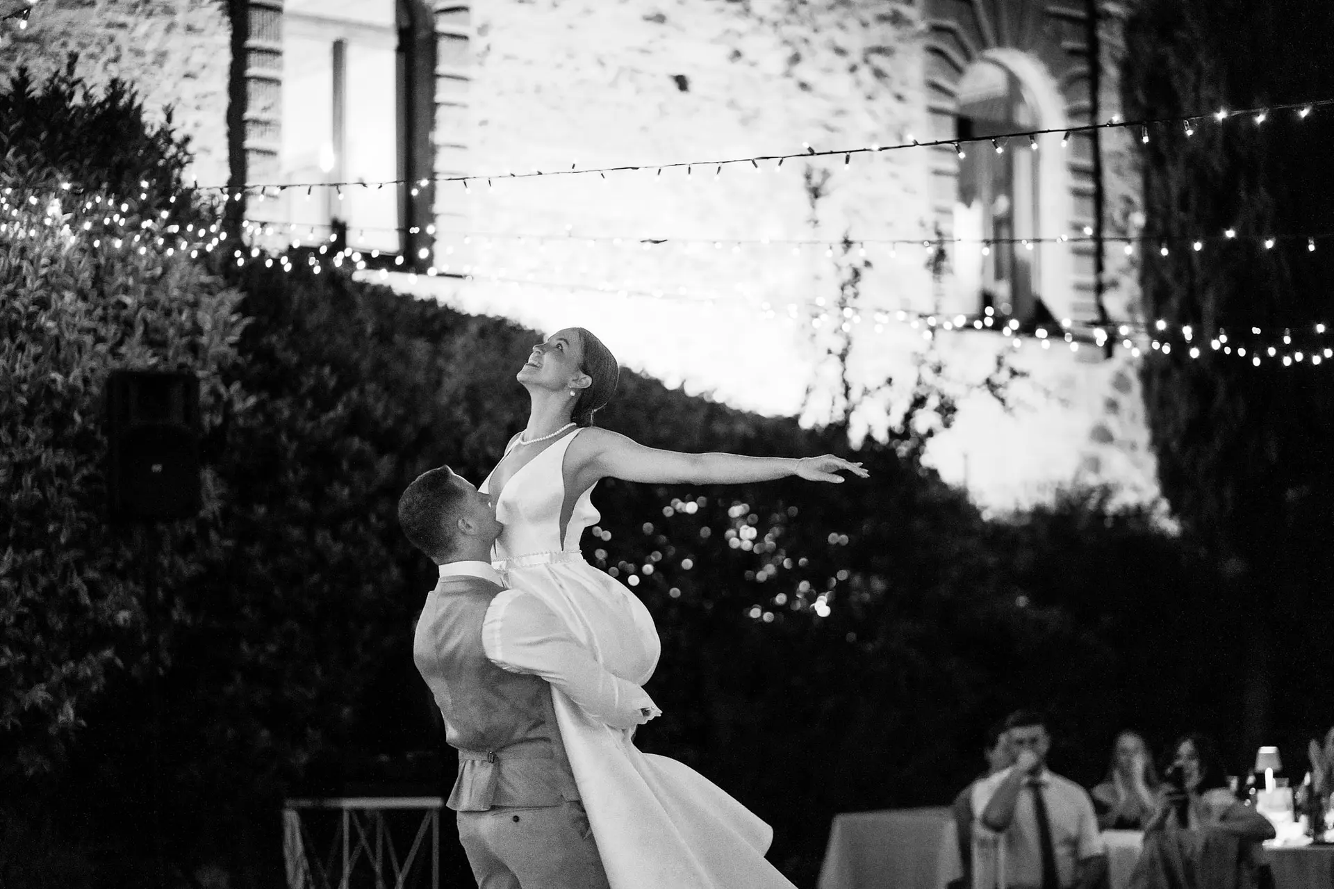 B&W: Groom joyfully lifts bride during their first dance to romantic Italian wedding music under lights.
