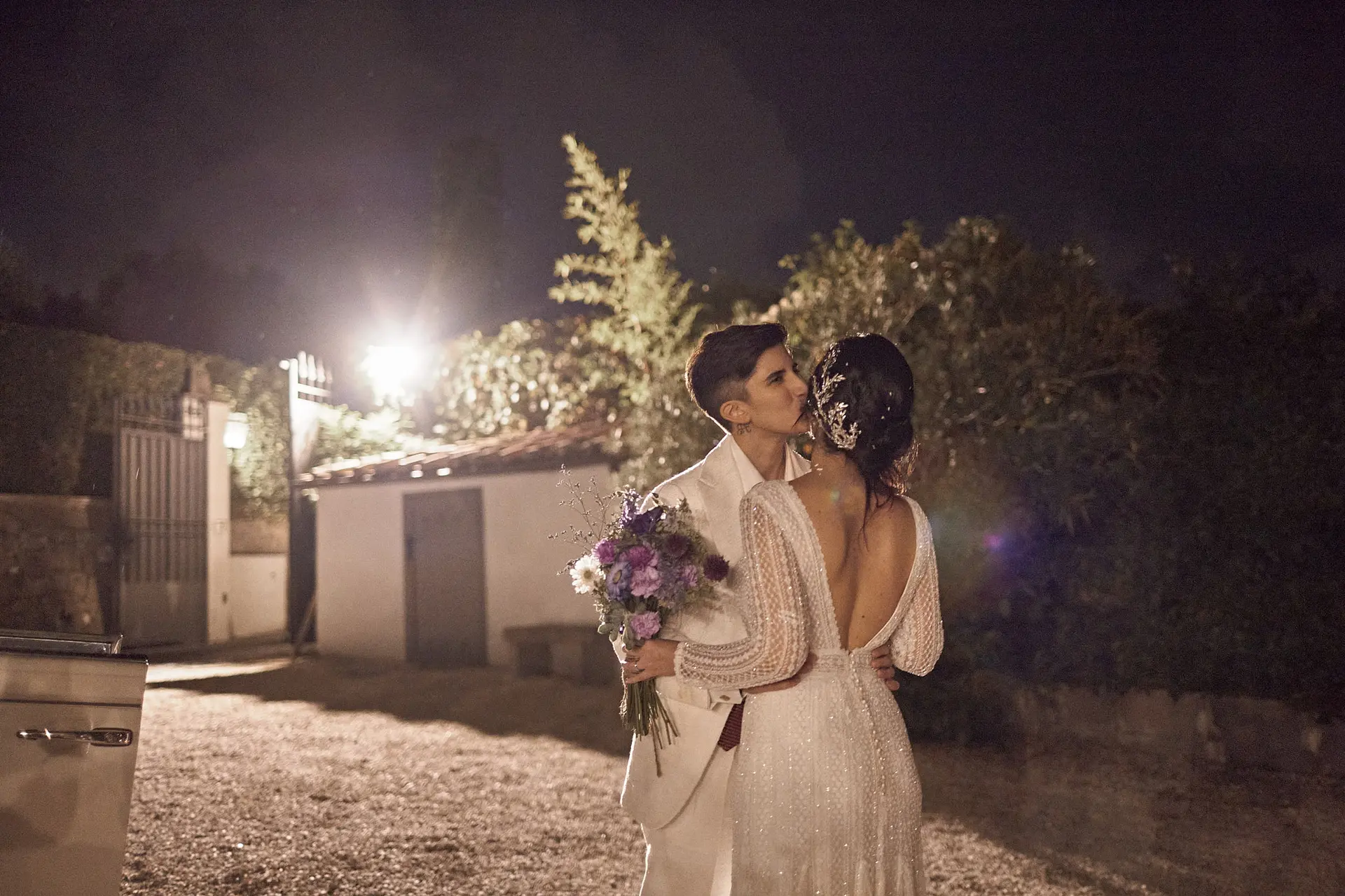 Two brides share a romantic night kiss outdoors; one holds a purple bouquet, the other wears a white suit.