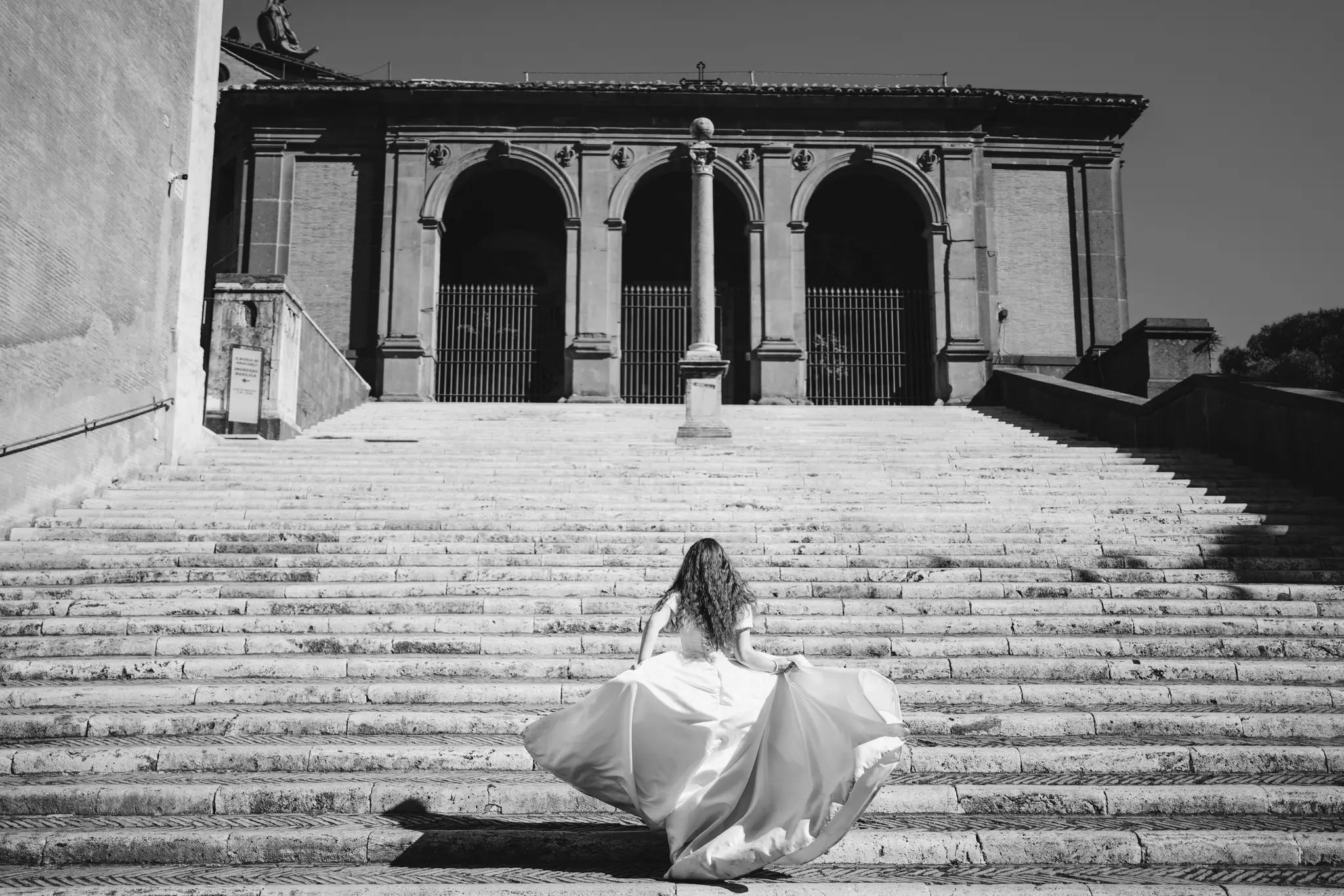 B&W: Bride in flowing wedding gown runs up grand historical stone steps towards arched building.