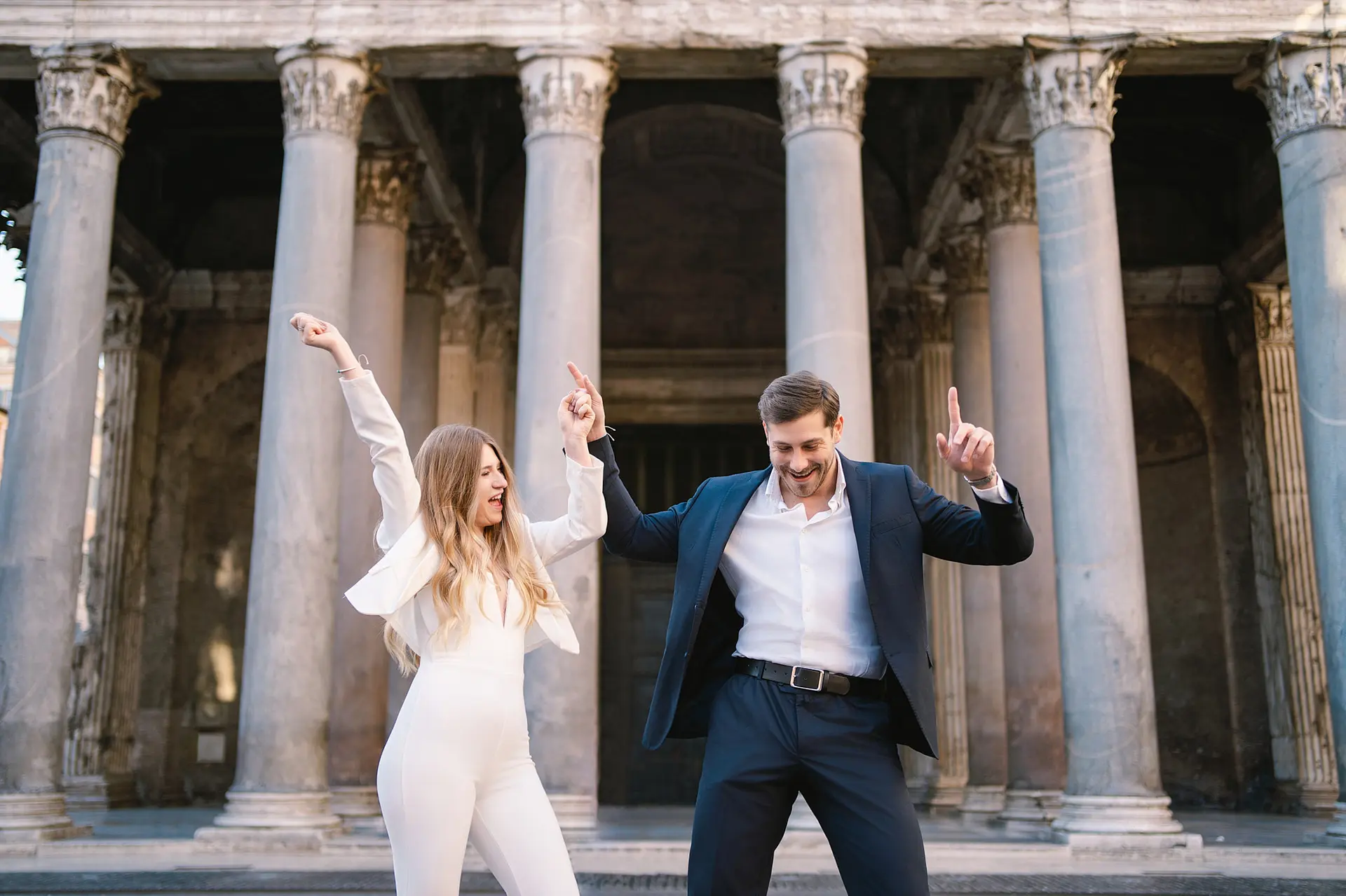 Joyful couple in stylish outfits dances energetically outdoors before Rome's Pantheon columns.