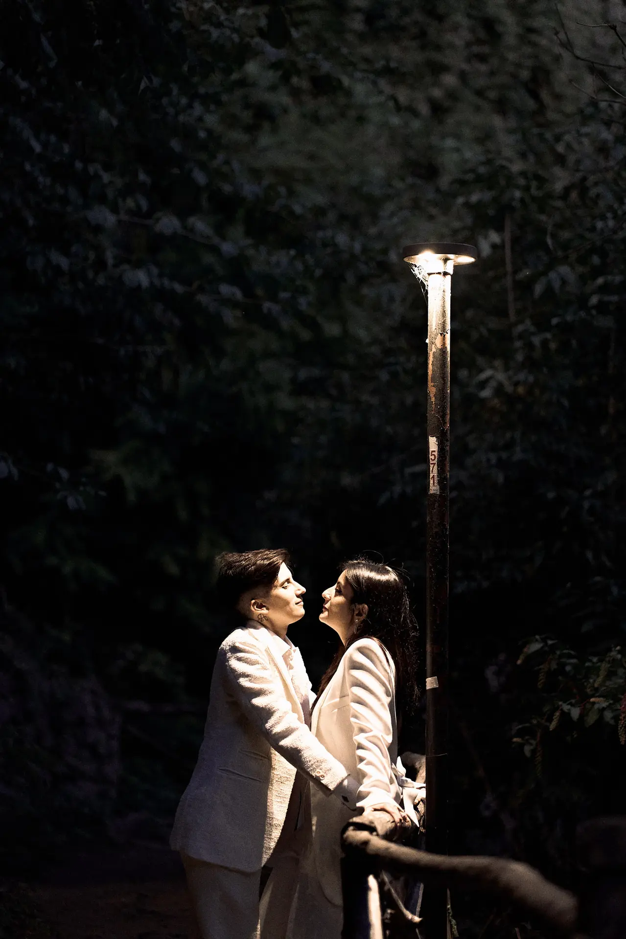 Two women in white suits gaze at each other under a lamppost after getting engaged.