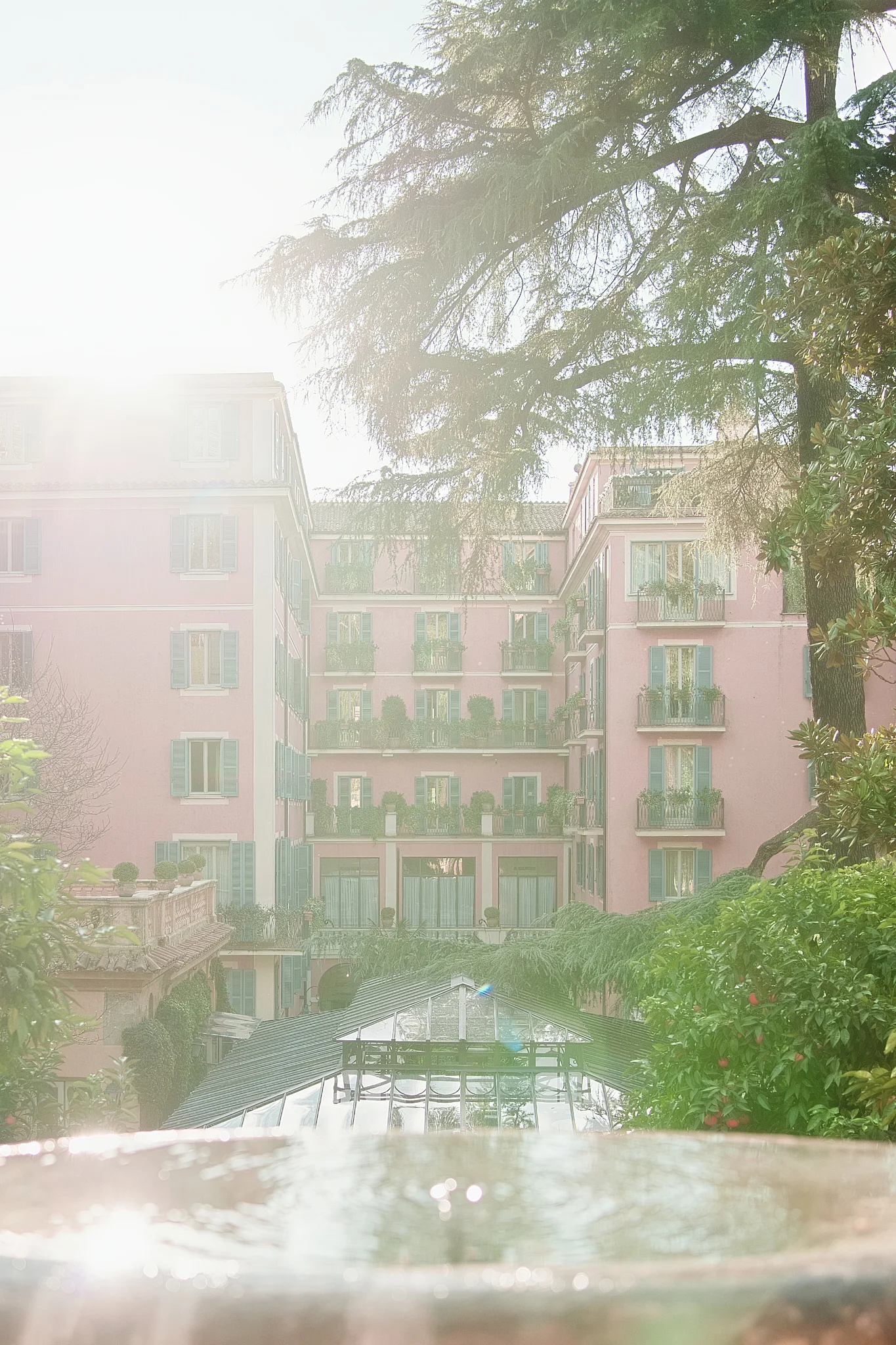 Sunlit view of the pink Hotel De Russie in Rome, with a fountain in the foreground.