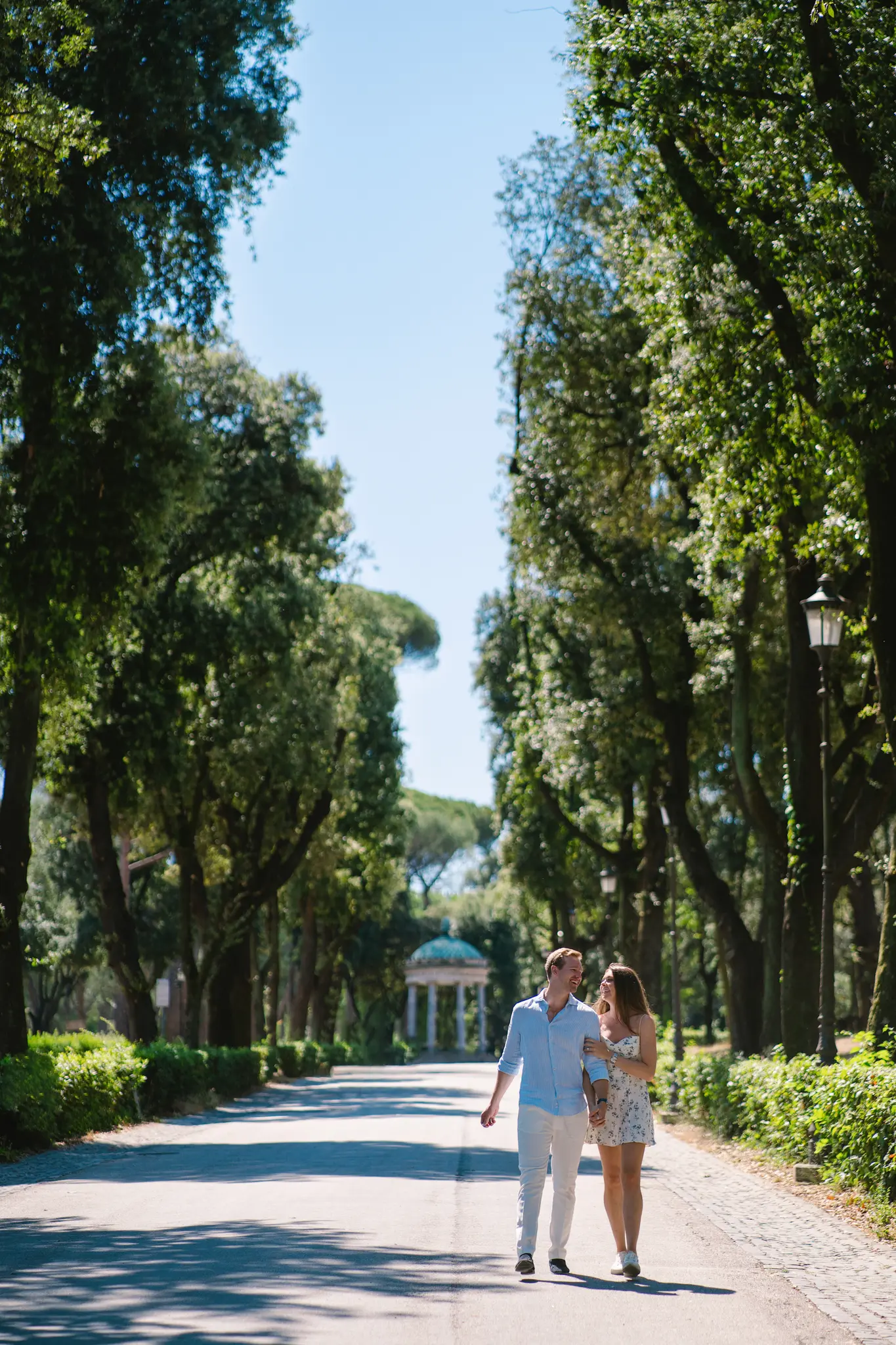 A couple strolls arm-in-arm down a sun-dappled, tree-lined avenue, a romantic park engagement photo.