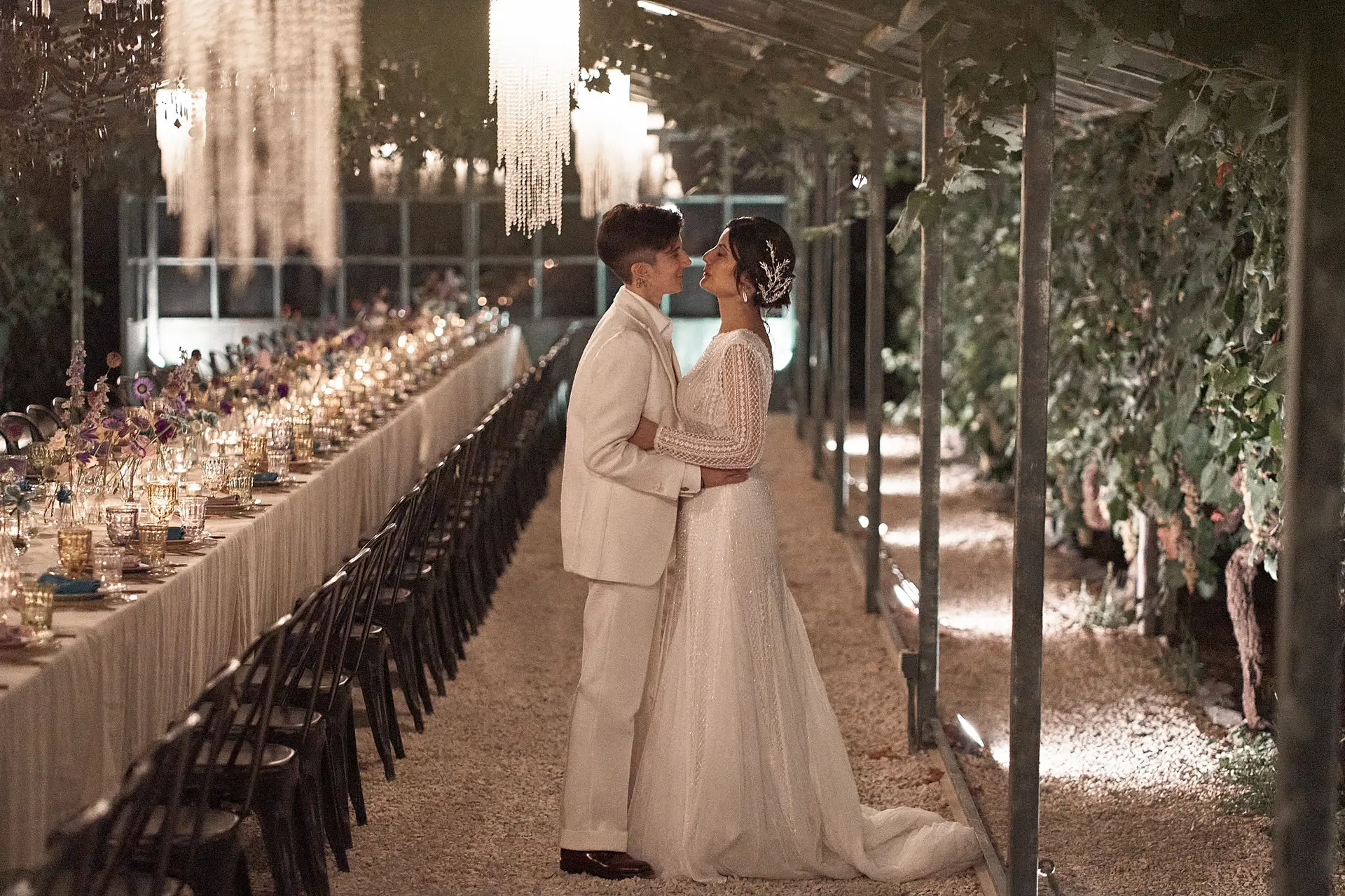 Two brides, one in a wedding gown and one in a white suit, embrace at their outdoor evening reception.