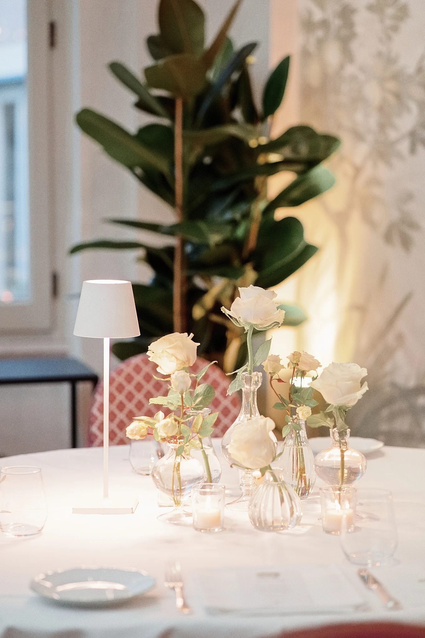 Close-up of a wedding table with white roses in small vases, candles, and a modern lamp. Preparing questions to ask a wedding vendor helps clarify floral options.