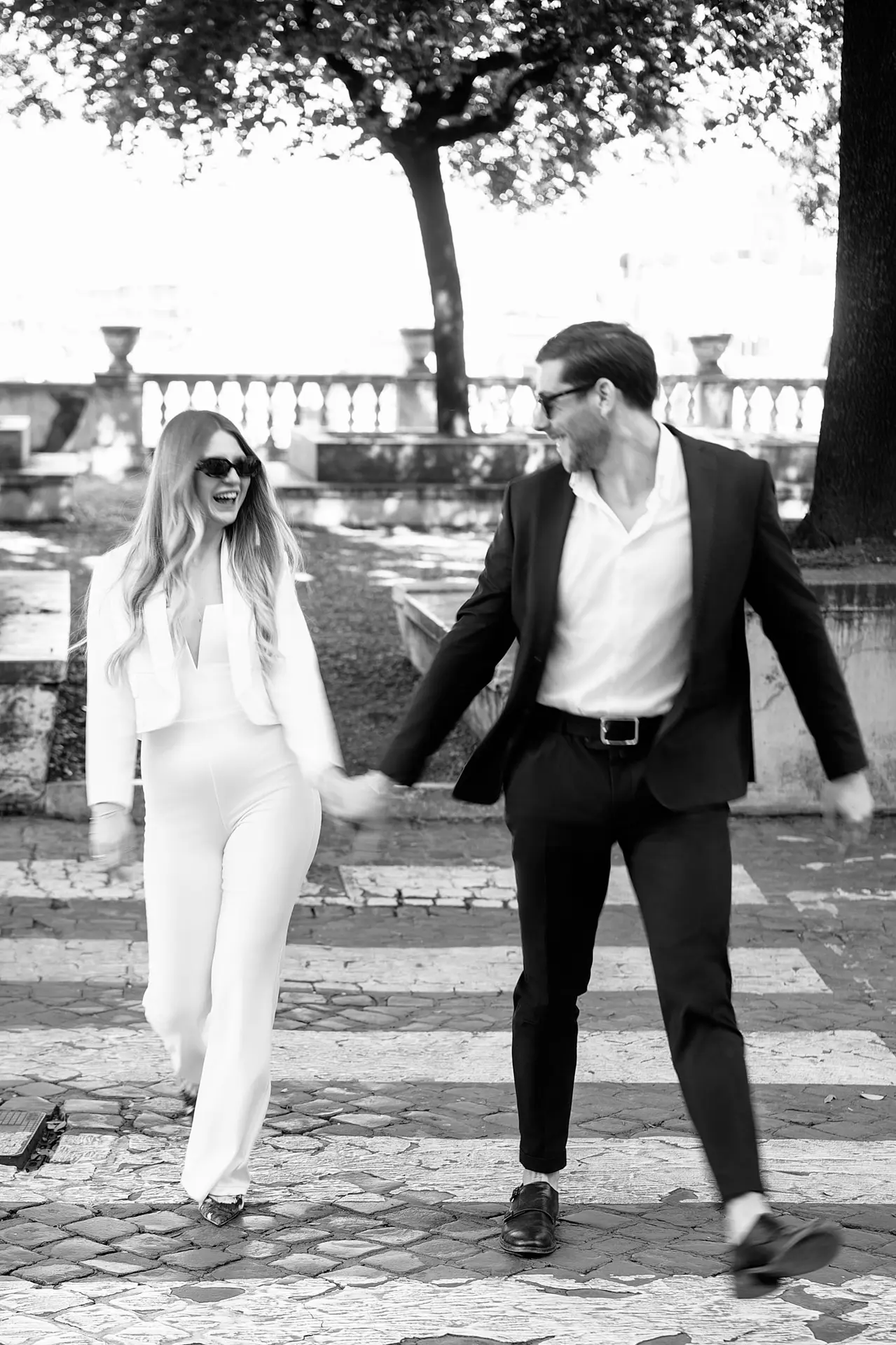 A joyful couple laughs while walking hand-in-hand. The bride wears a stylish white jumpsuit.