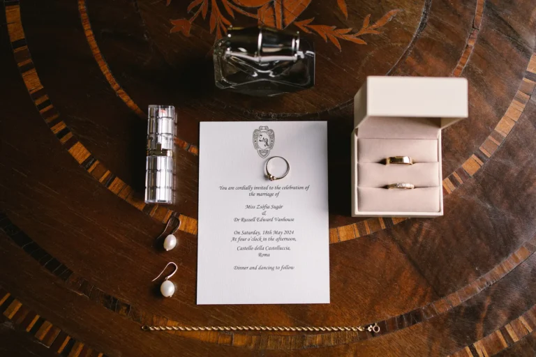 A classic wedding invitation flat lay with rings, lipstick, and perfume on a wooden table.