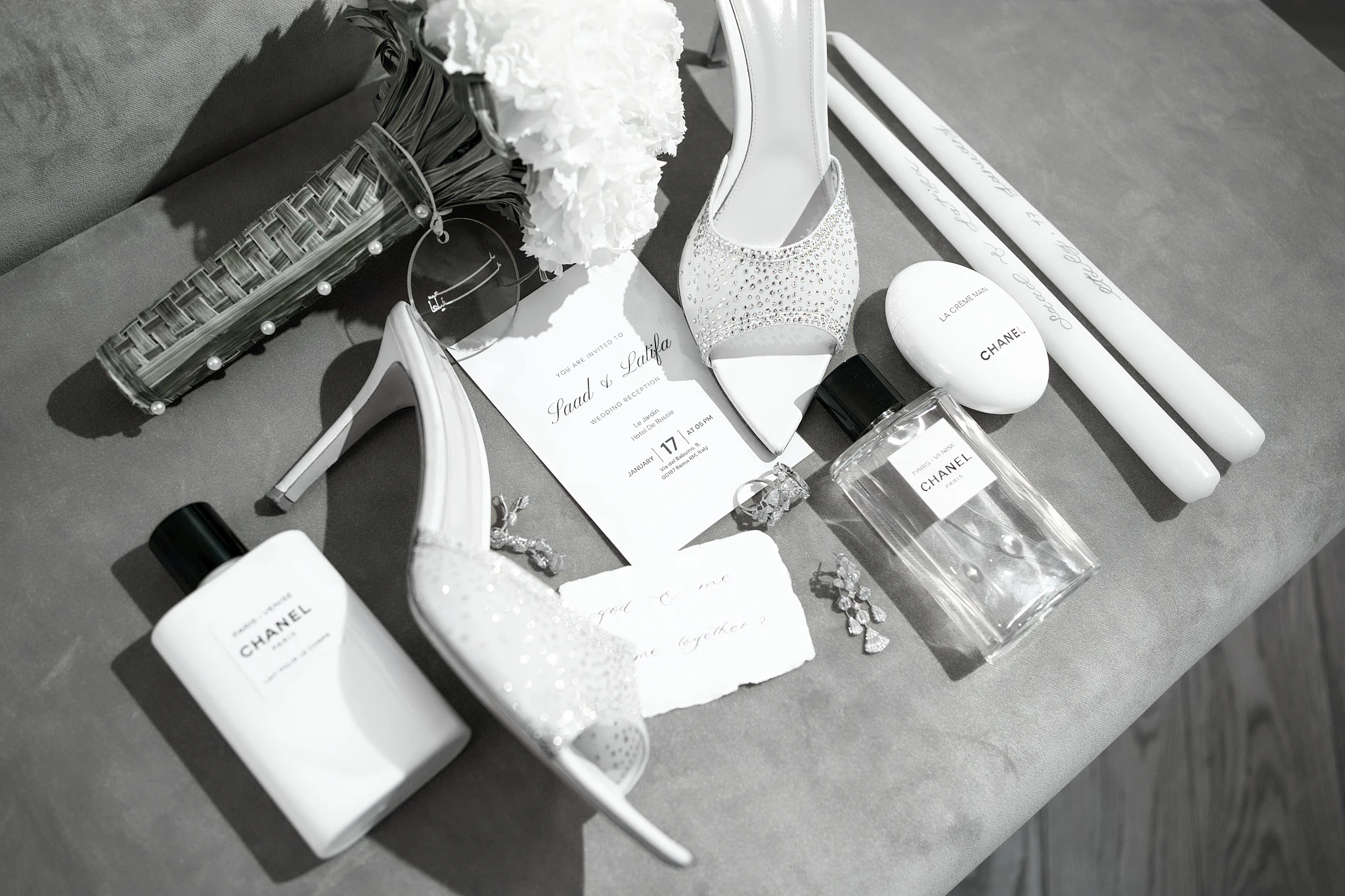 A black and white flat lay of wedding details: Chanel perfume, sparkling heels, and a bouquet.