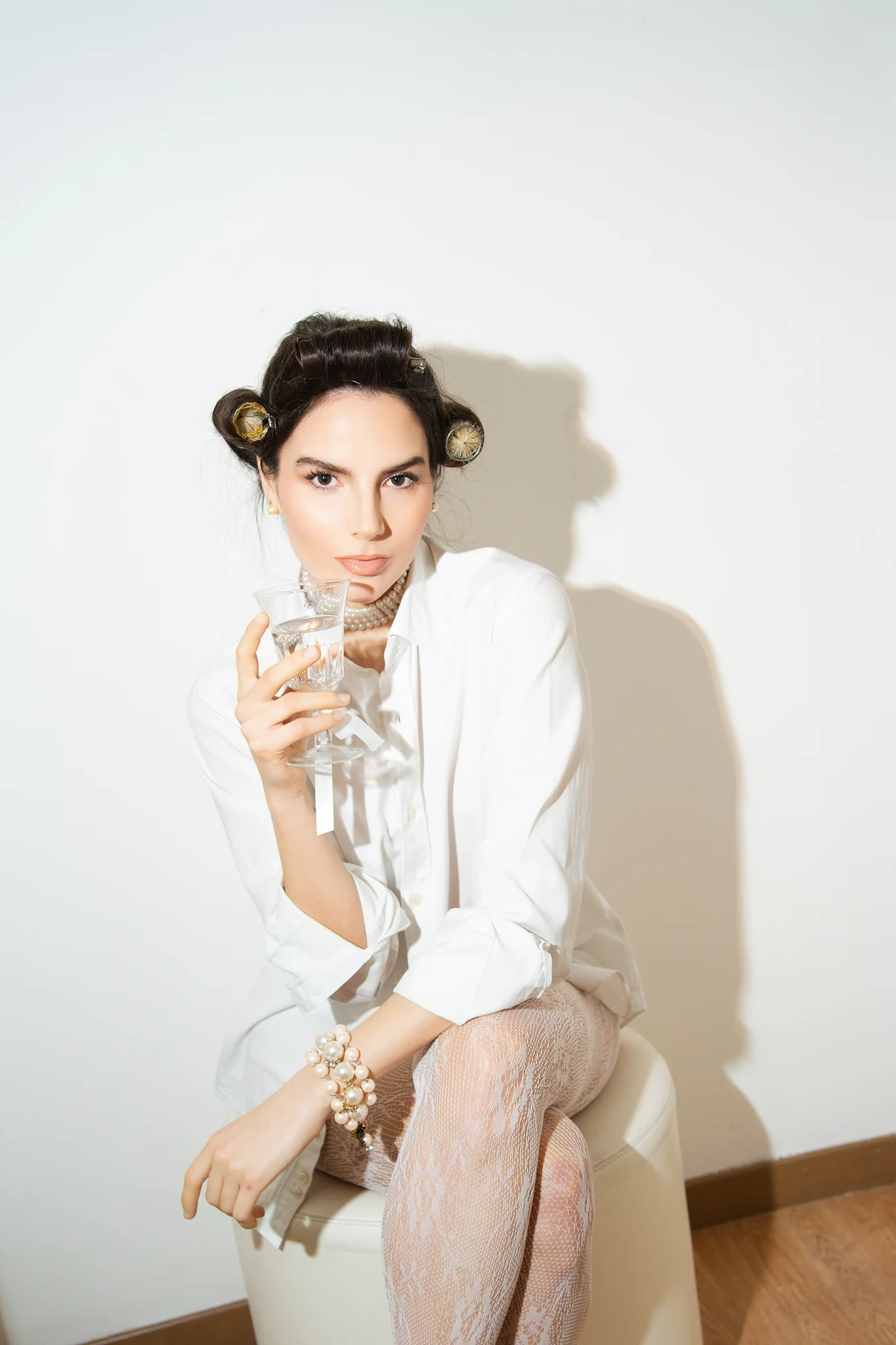 A chic bride-to-be with hair in curlers, wearing a white shirt and pearls, holds a glass.