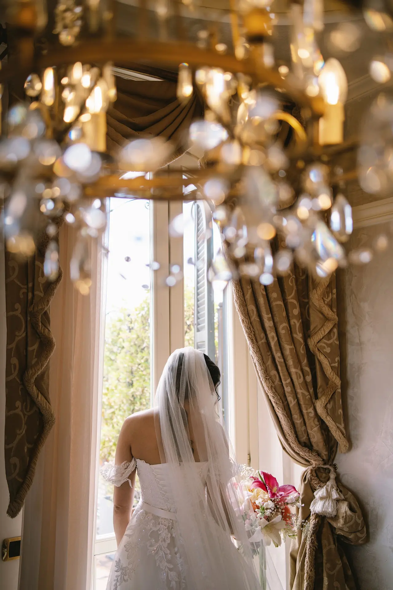 A bride in her gown and veil looks out a window, framed by a beautiful crystal chandelier.