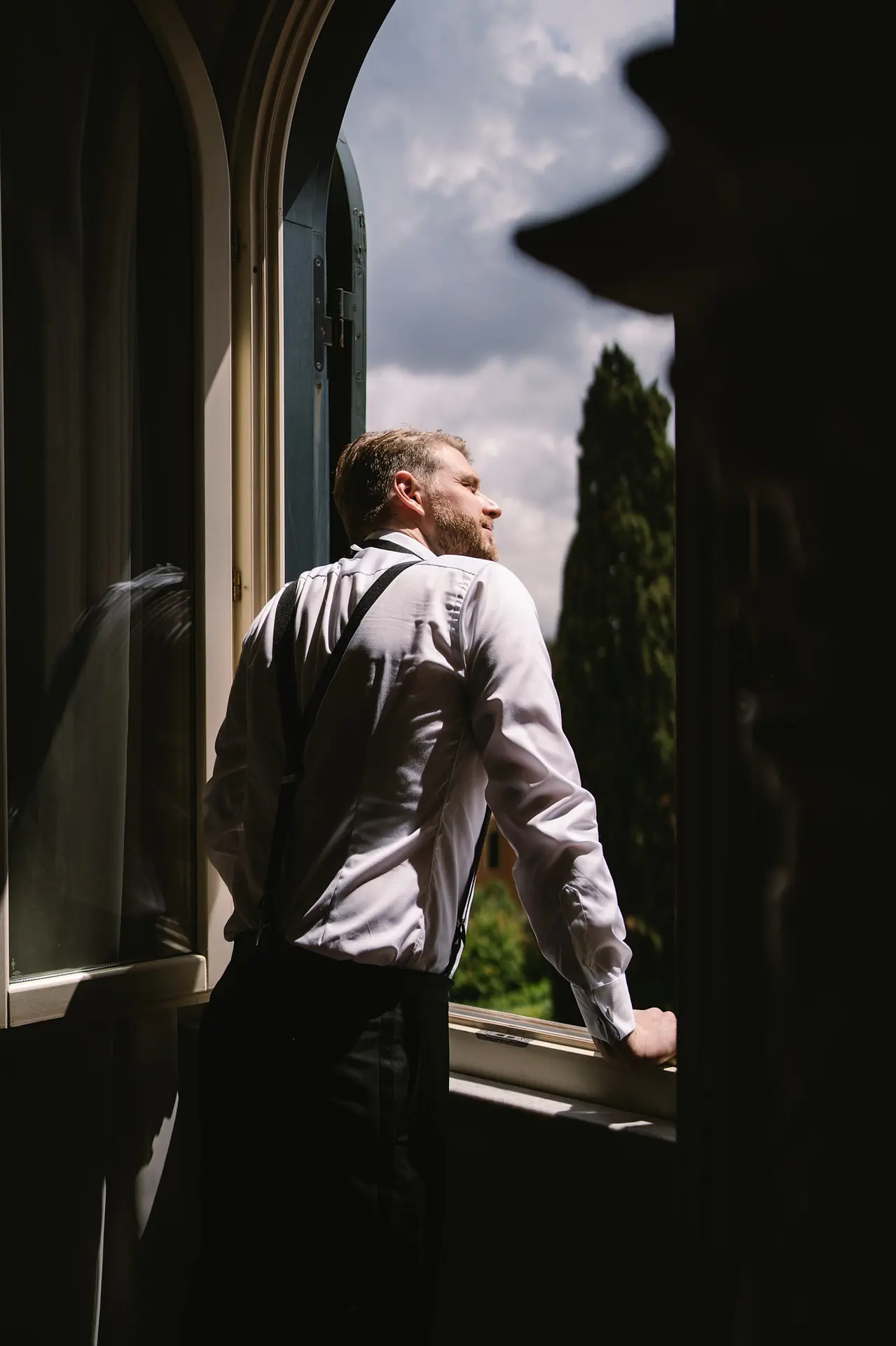 A groom in a white shirt and suspenders thoughtfully looks out an open window into the sun.