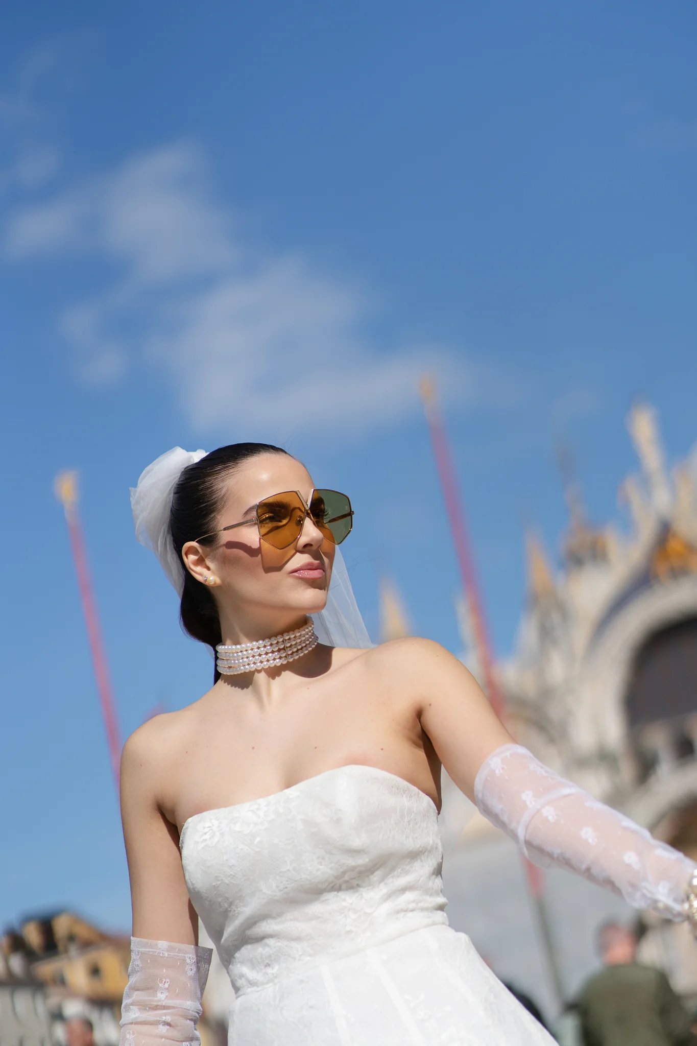 A modern bride in a strapless gown, short veil, and sunglasses against a bright blue sky.