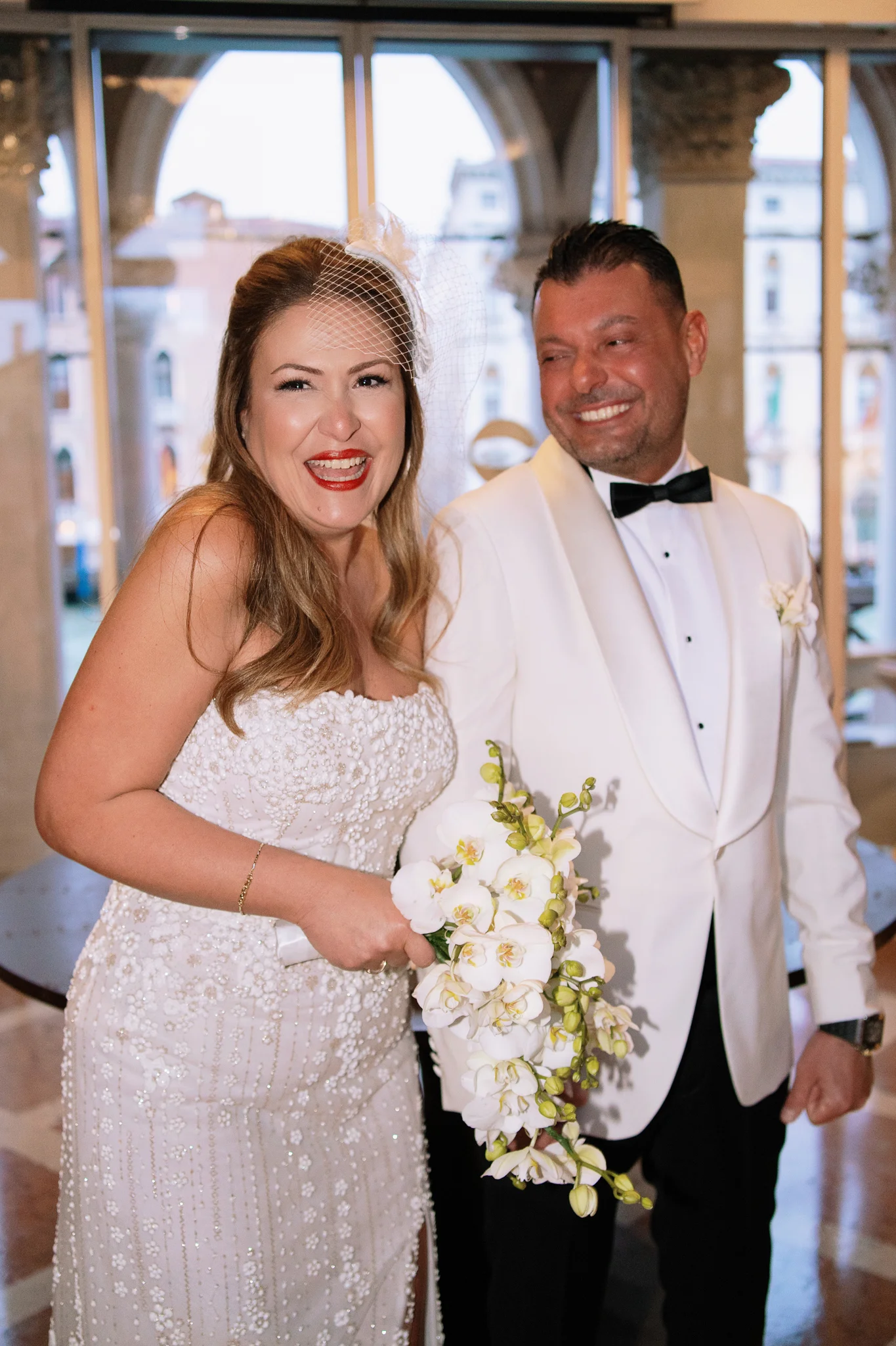A joyful bride in a birdcage veil laughs next to her groom in a white tuxedo jacket during their wedding.