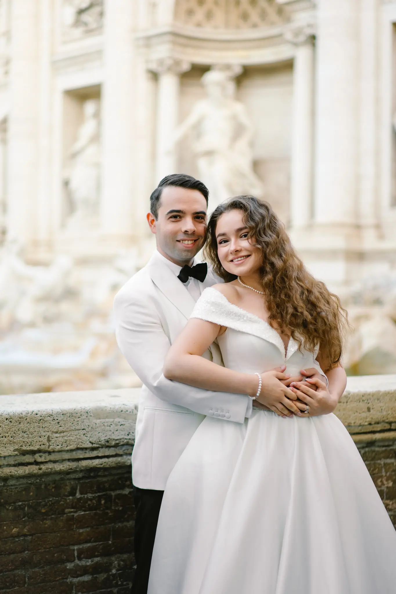 A groom in a white tuxedo jacket embraces his bride in front of a classic Italian fountain.