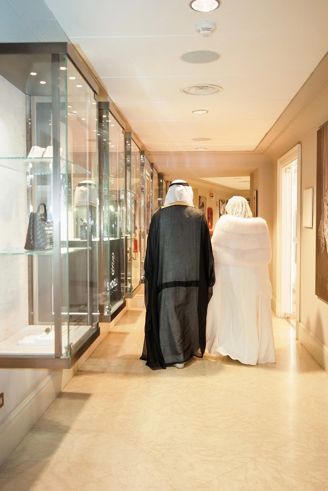 A bride and groom in traditional Arabic wedding attire walk down a hall lined with glass display cases.