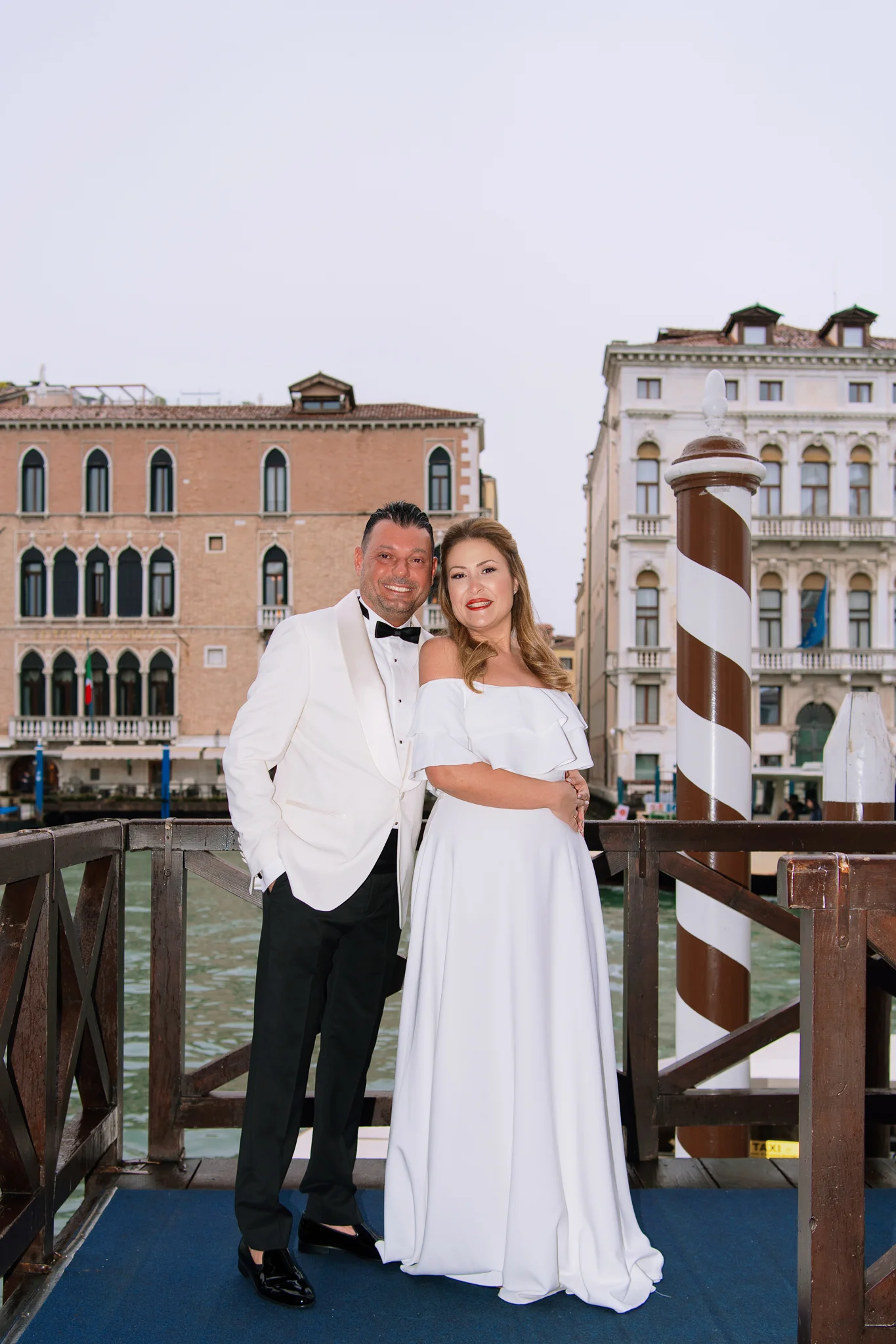 A happy couple poses on a dock in Venice, with a canal and traditional Venetian architecture behind them.