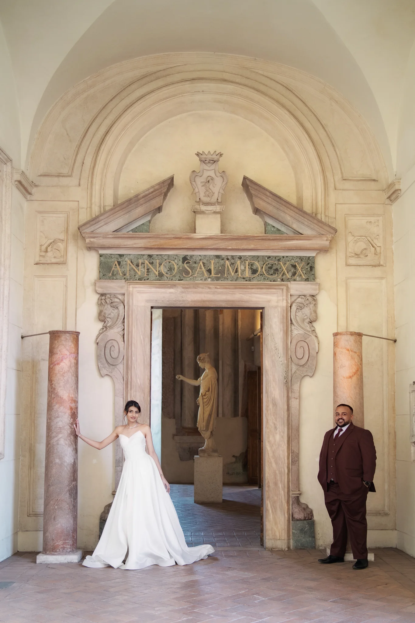 A bride and groom pose elegantly in a grand Italian archway with a statue visible in the background.