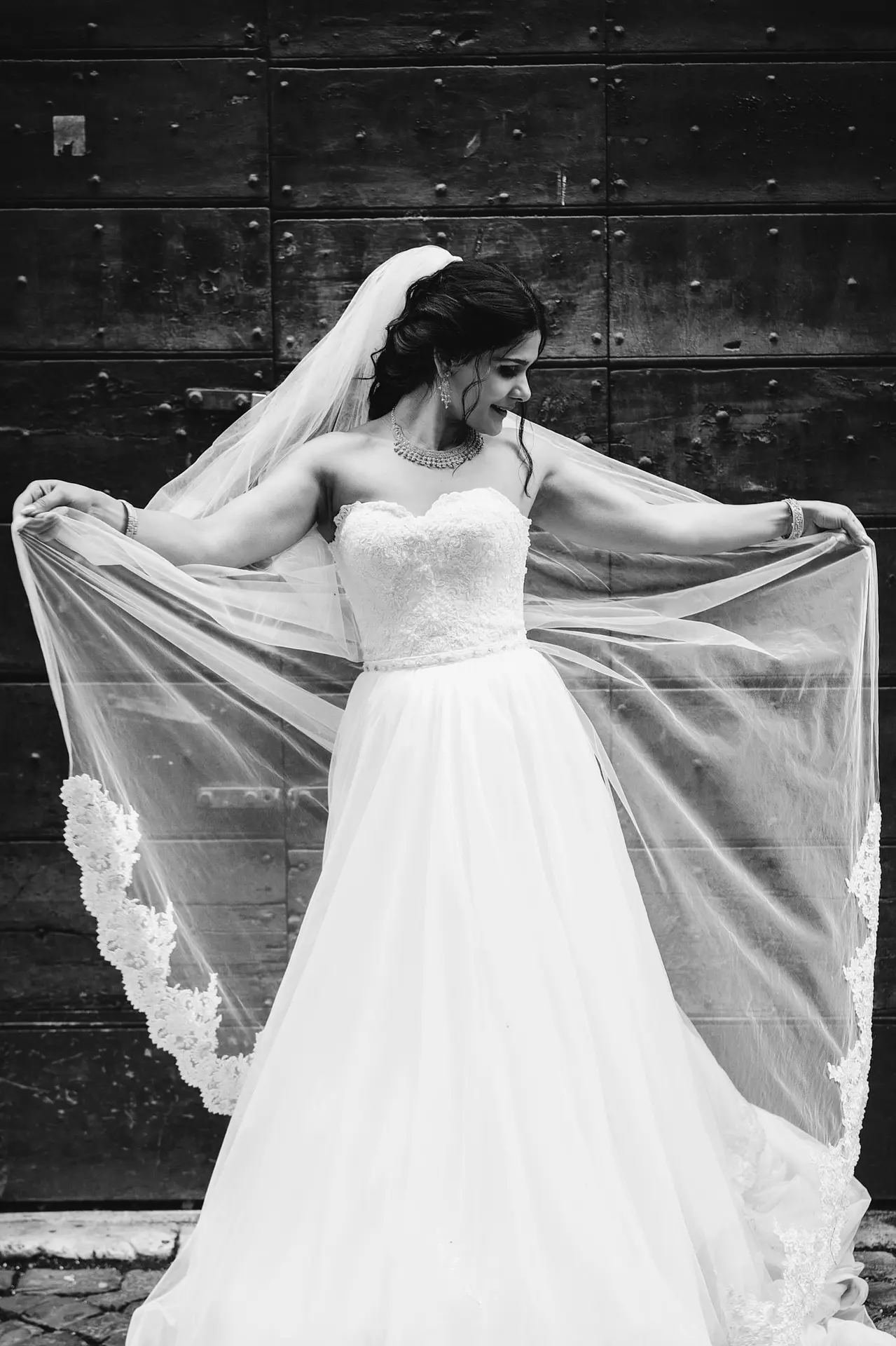 A beautiful bride in a strapless gown smiles while holding out her long, elegant veil.