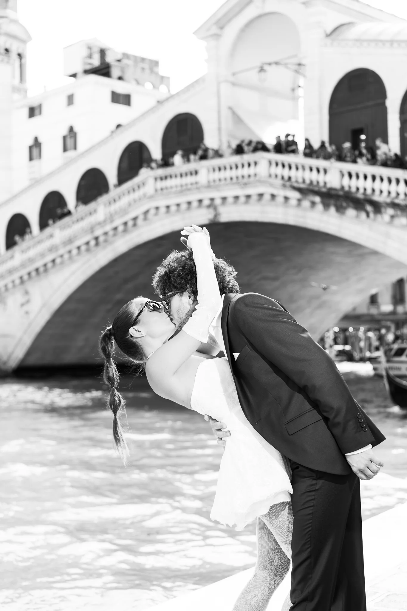 A couple shares a passionate kiss in black and white, with the Rialto Bridge in Venice behind them.