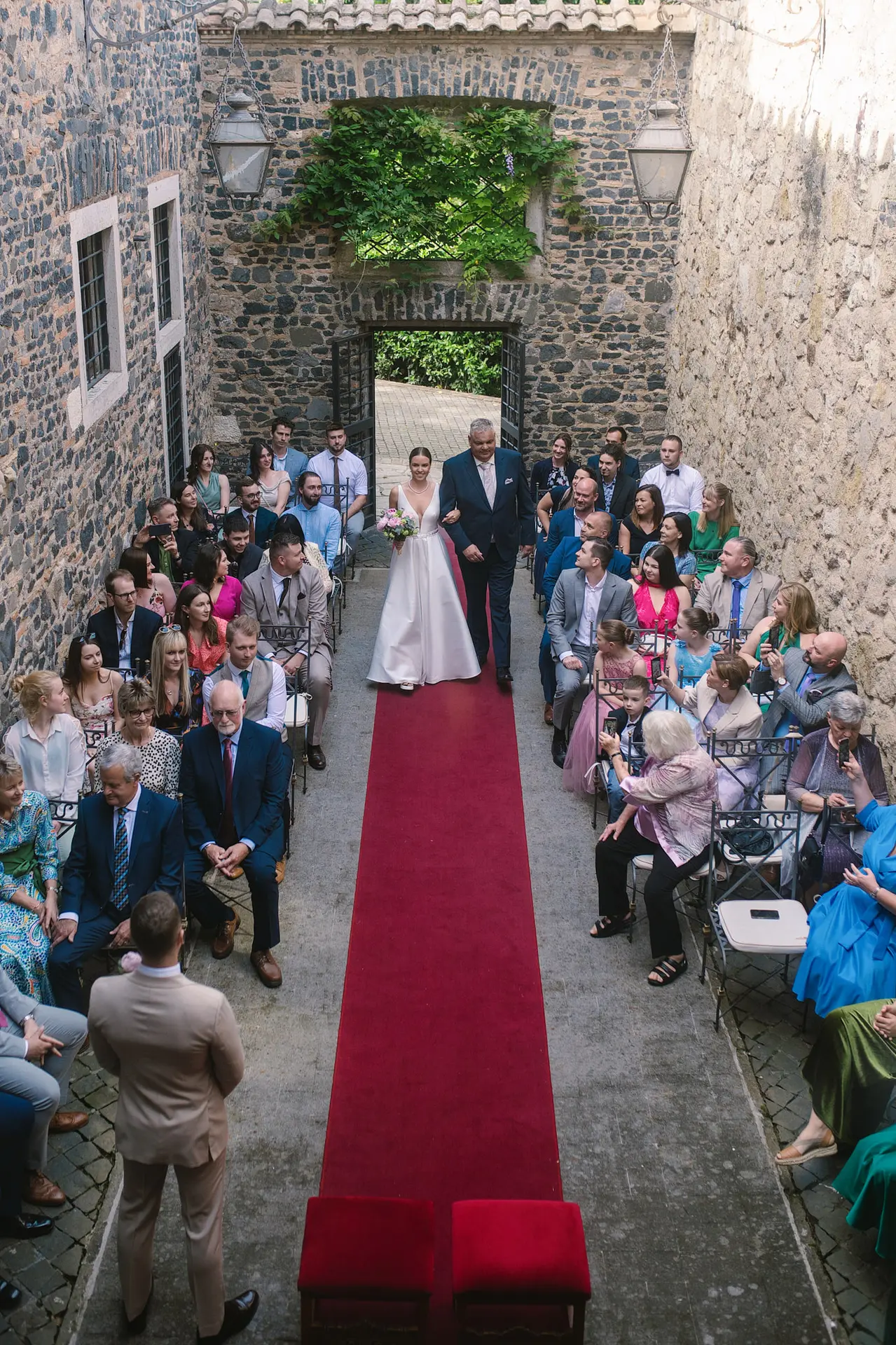 A bride is walked down a long red aisle by her father in a beautiful stone courtyard ceremony.