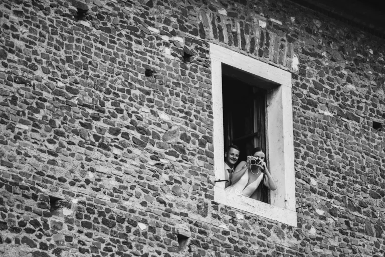 Two newlyweds peek from a window in an old stone building, one holding a vintage camera. B&W photo.