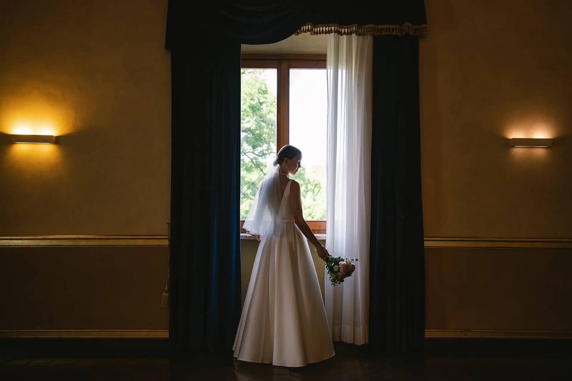 A bride in her wedding dress stands in silhouette, holding her bouquet while looking out a large window.