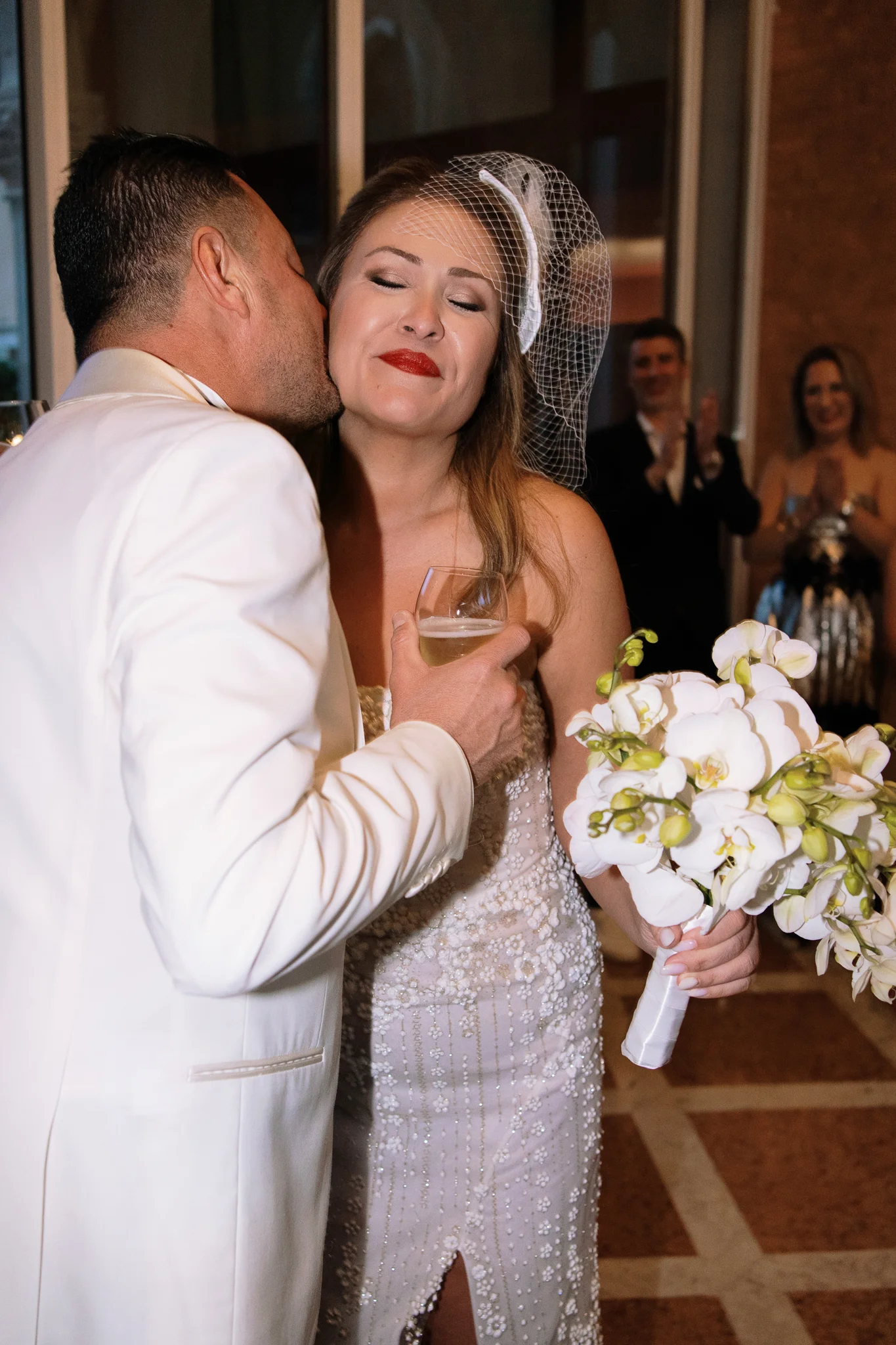 A groom kisses his smiling bride on the cheek. They are holding a glass of champagne and an orchid bouquet.