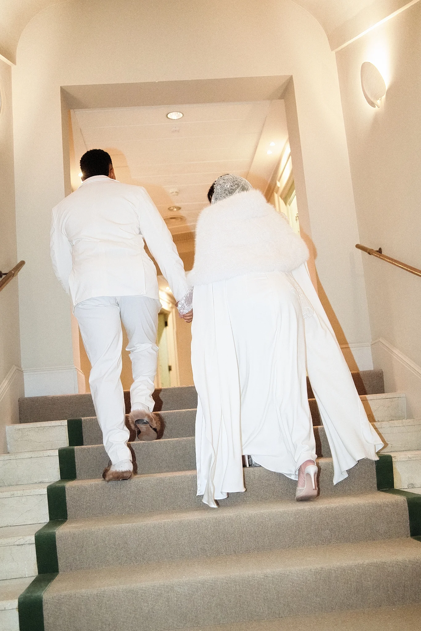 From behind, a bride and groom in stylish all-white outfits walk hand-in-hand up a staircase.