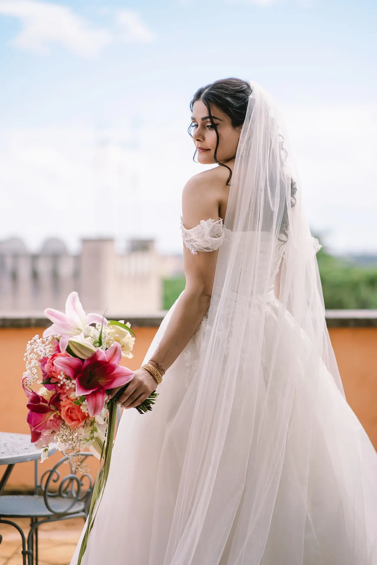 A beautiful bride with a long veil stands on a balcony, looking over her shoulder holding her bouquet.