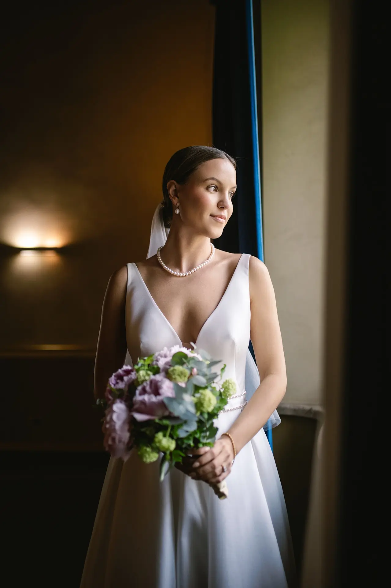 An elegant bride in a satin V-neck gown and pearls holds her bouquet, looking out a window.