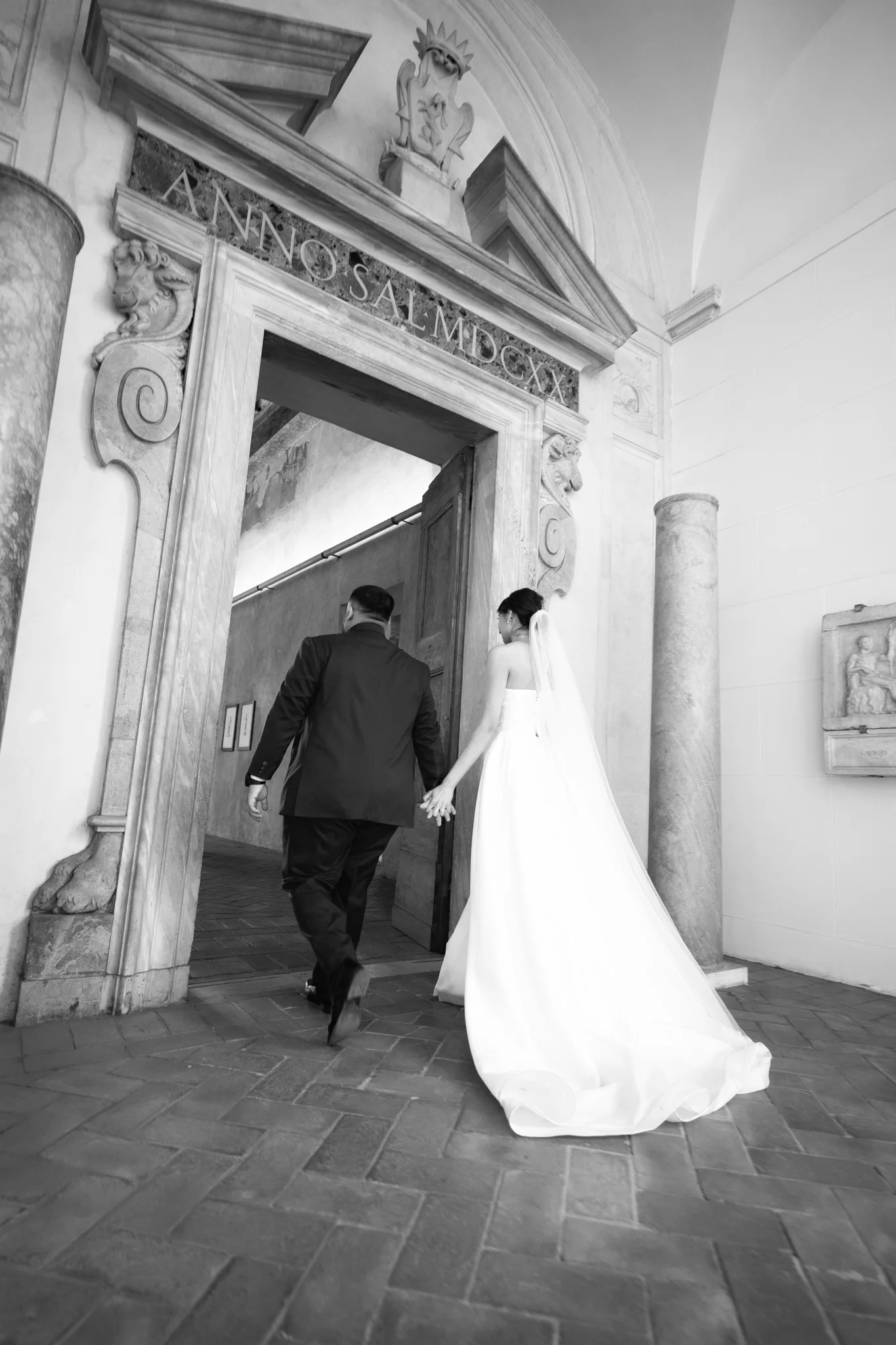 Black and white photo of a bride and groom holding hands as they walk through an ornate, historic doorway.