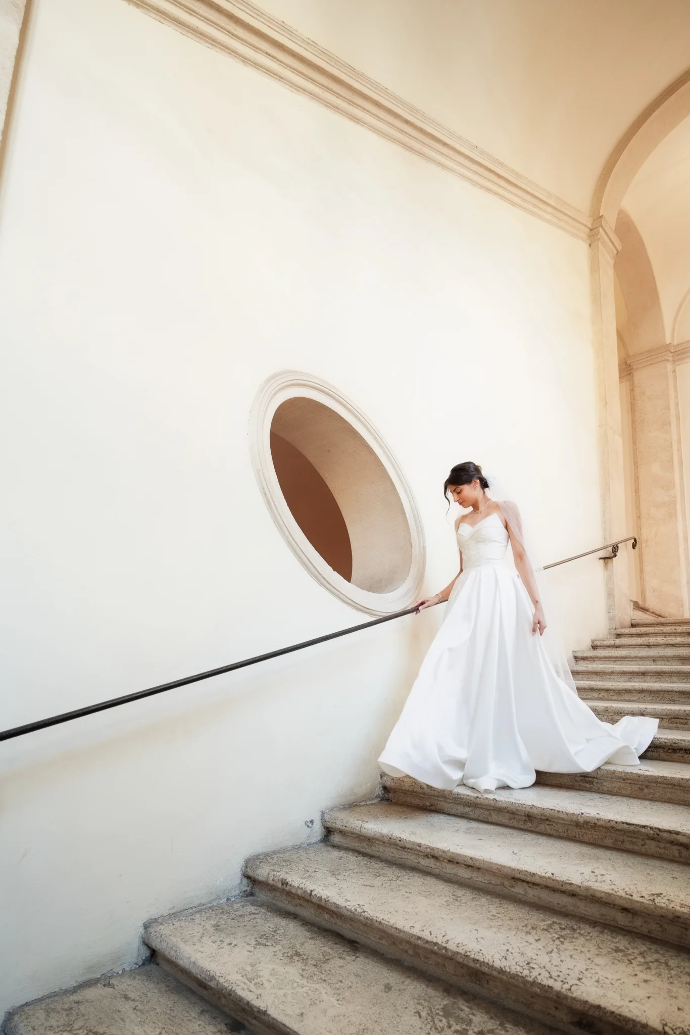 A bride in a flowing white gown descends a grand stone staircase, holding the handrail in a classic building.