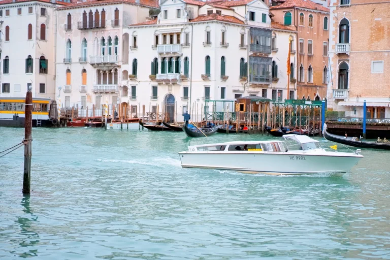 A white water taxi cruises along the Grand Canal in Venice, with historic buildings in the background.