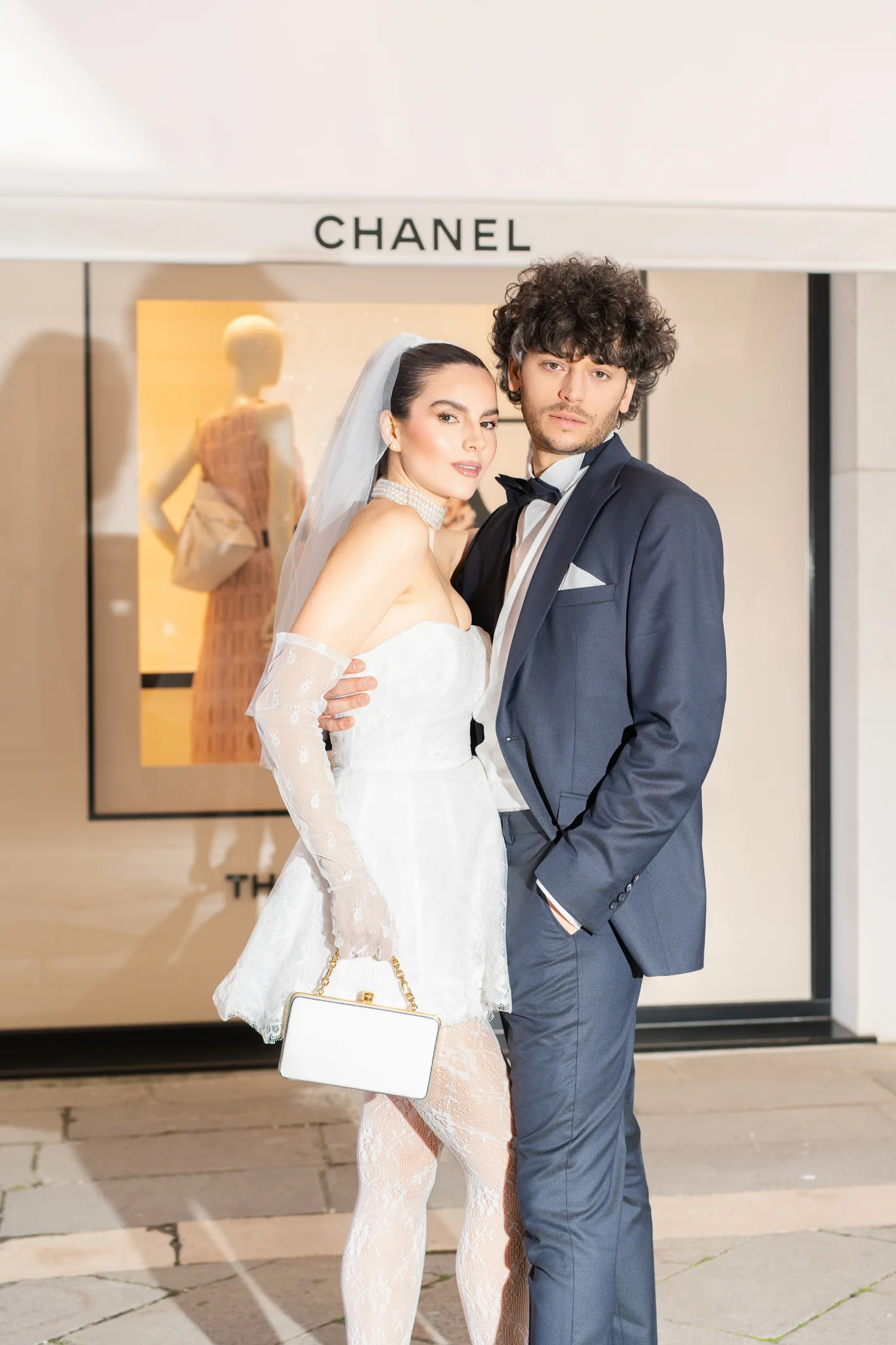 A chic bride and groom in modern wedding attire pose for a photo in front of a Chanel store.