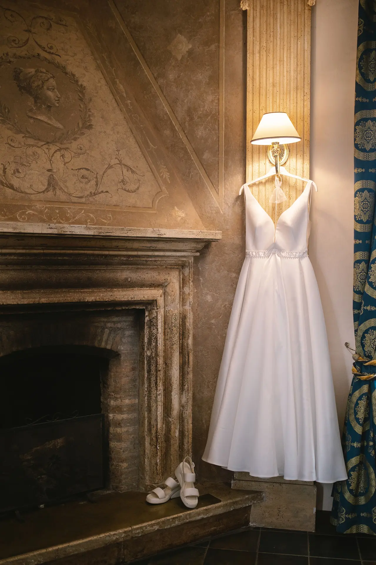 A timeless white wedding dress hangs from a sconce next to a classic stone fireplace in Italy.
