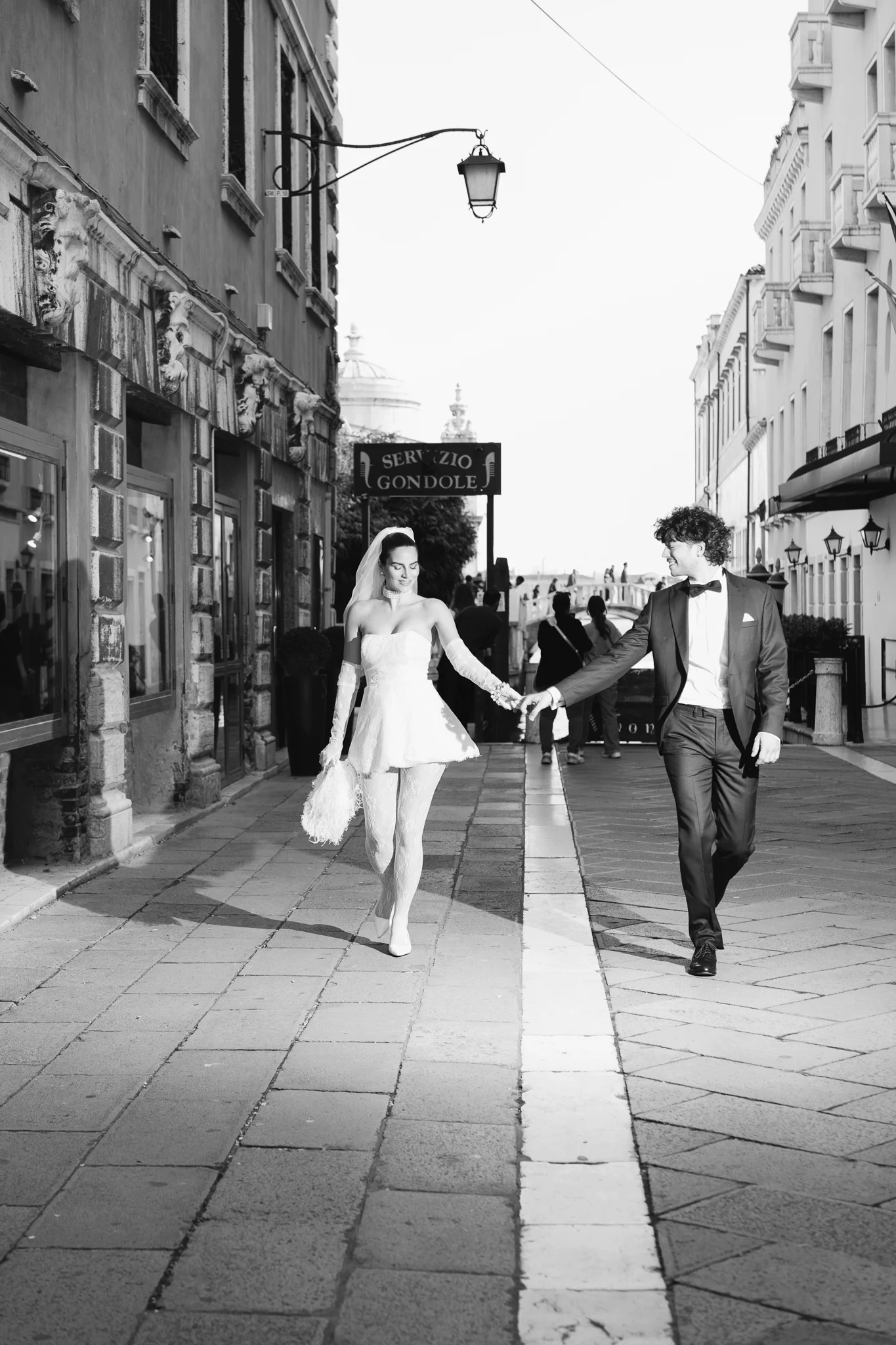 B&W: A bride and groom walk hand-in-hand down a narrow cobblestone street in Venice, Italy.