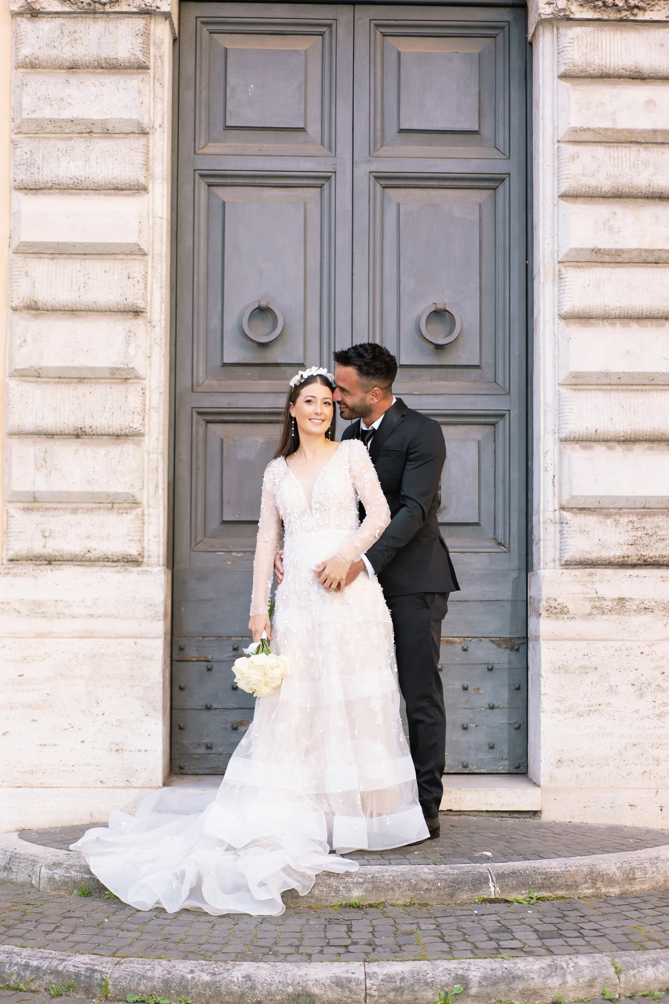 An elegant groom embraces his bride, who holds white roses, in front of a grand, classic door.