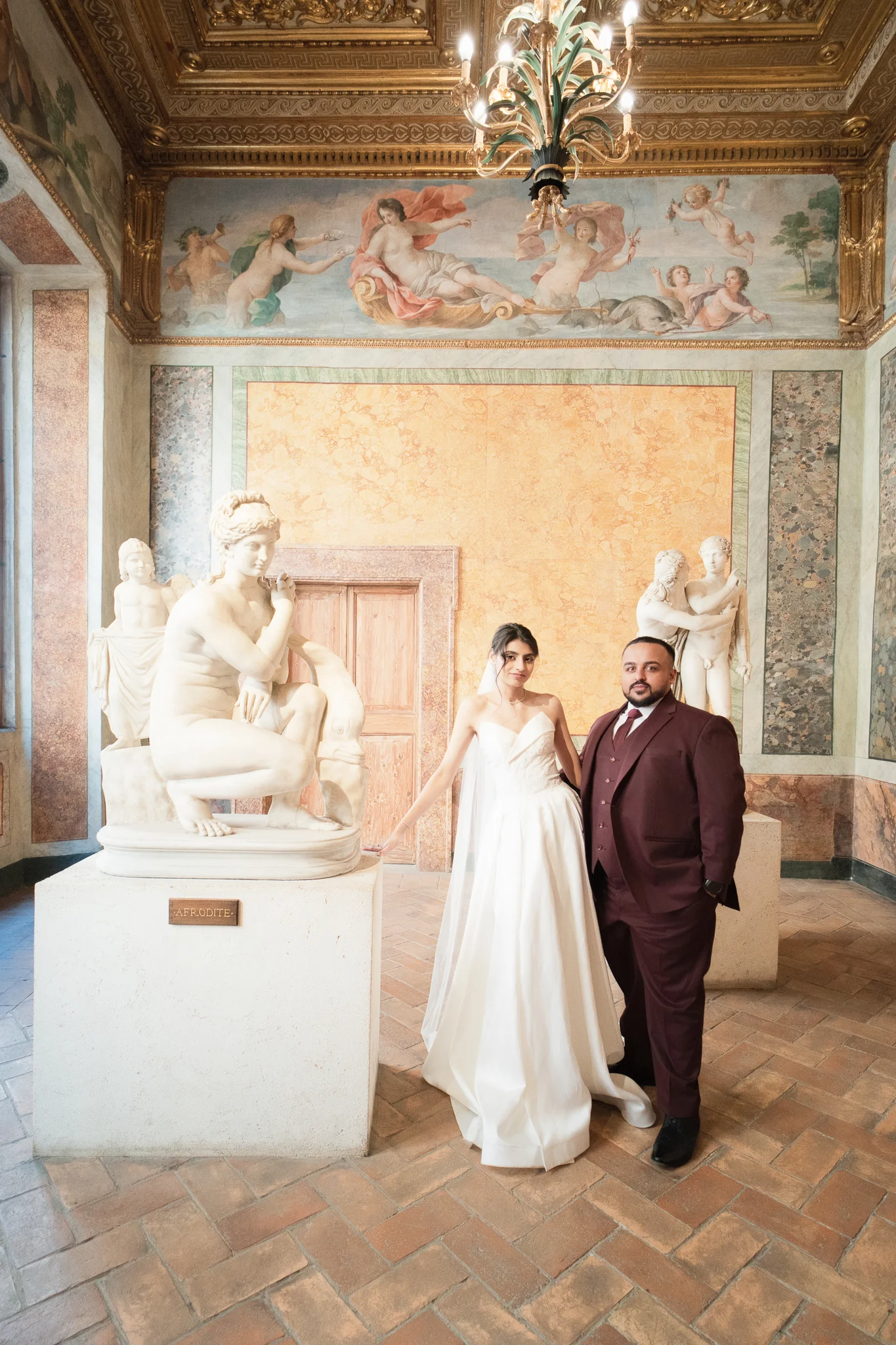 A couple poses with statues in an ornate hall. Venue choice affects the wedding photographer cost in Italy.