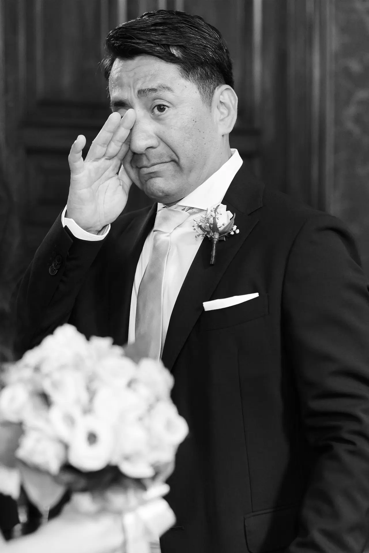 A groom becomes emotional and wipes a tear from his eye, captured in a poignant black and white photo.