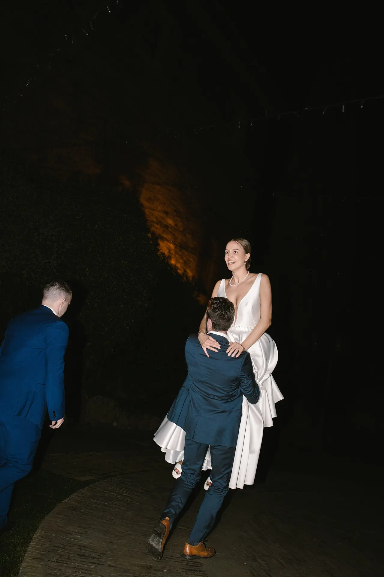 The bride in her white gown is playfully lifted by a guest during the outdoor evening celebrations.