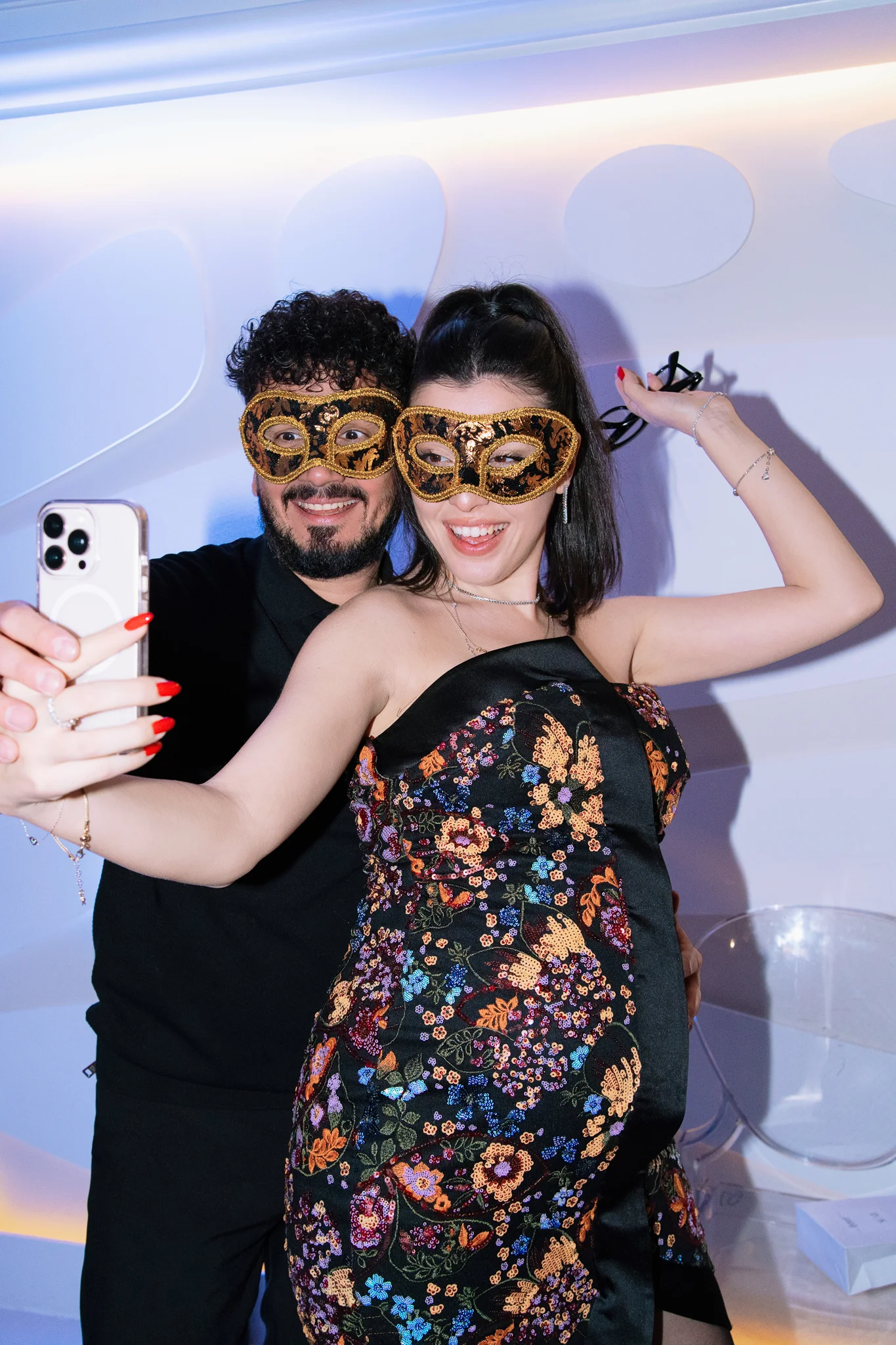 A happy couple wearing ornate gold and black masquerade masks take a fun selfie at a wedding party.
